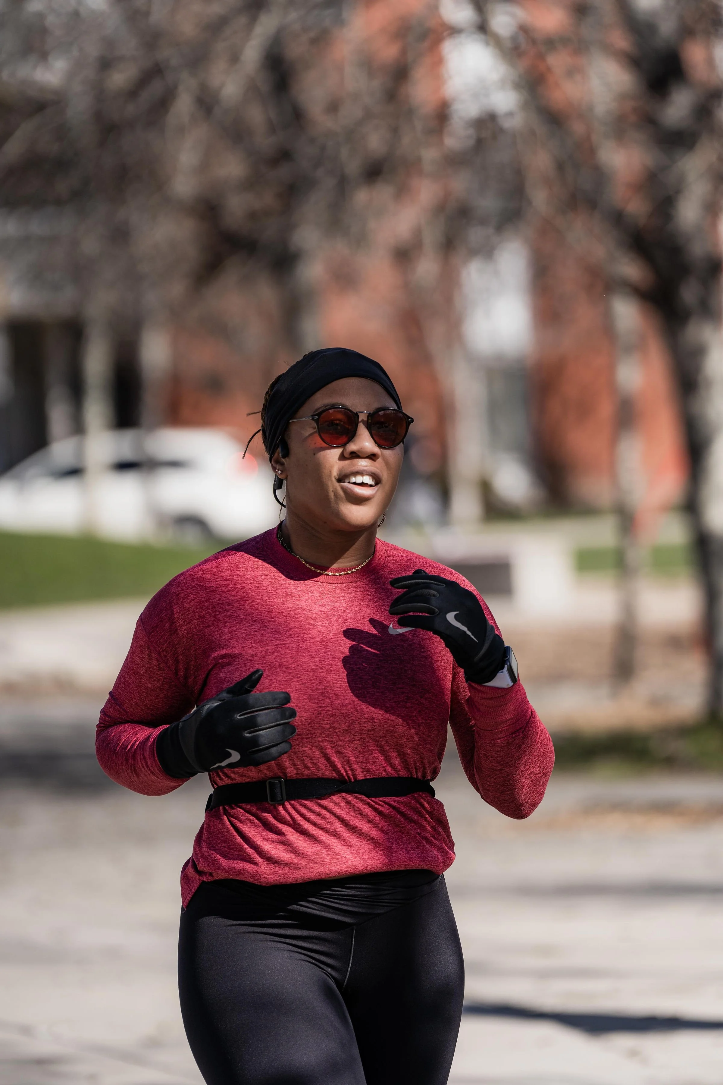 A woman running outdoors on a sunny day, wearing sunglasses, a black head covering, a red athletic long-sleeve shirt, black gloves, and black leggings.