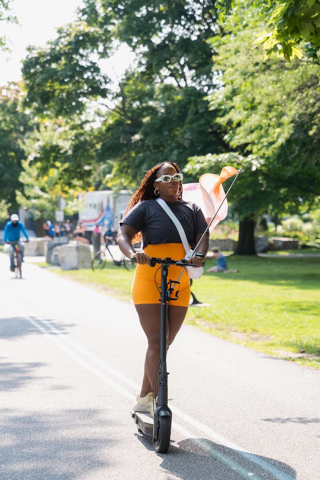 Woman riding an electric scooter on a sunny day in a park with green trees, holding a pink and orange flag.
