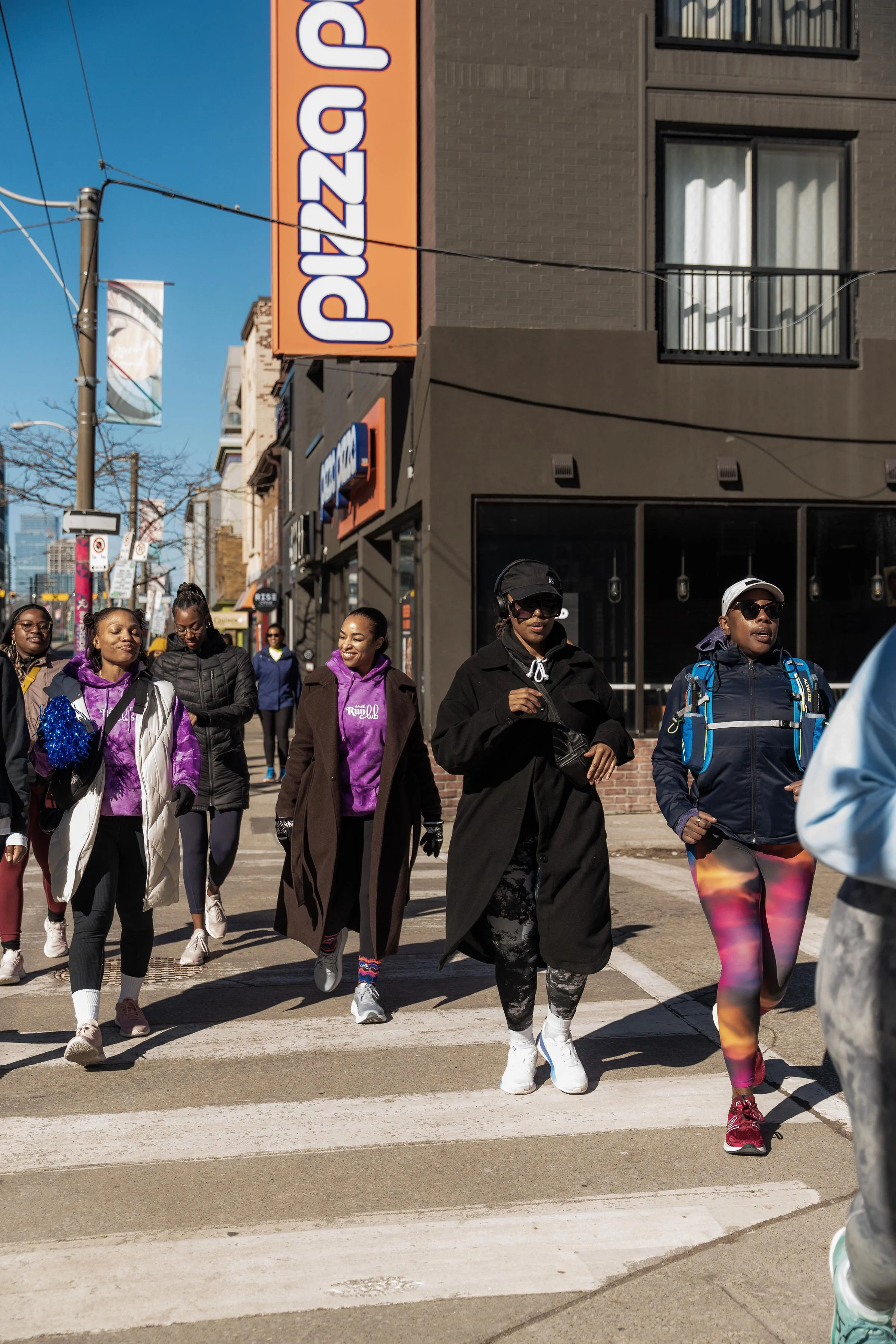 Group of women crossing the street in an urban setting, some wearing athletic or casual clothing, with a pizza restaurant sign visible on the building in the background.