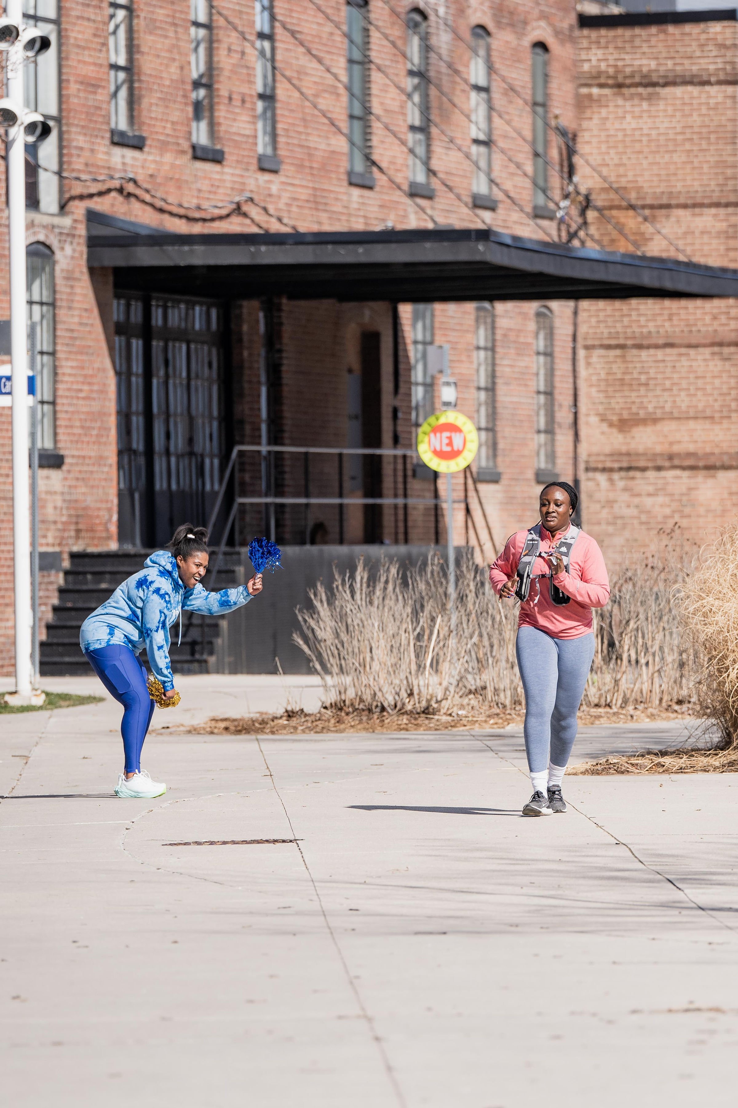 A girl in a blue hoodie and blue leggings cheerfully waving pom-poms as a woman in a pink hoodie and gray leggings walks on a sidewalk near a brick building, with dry bushes and a "NEW" sign in the background.