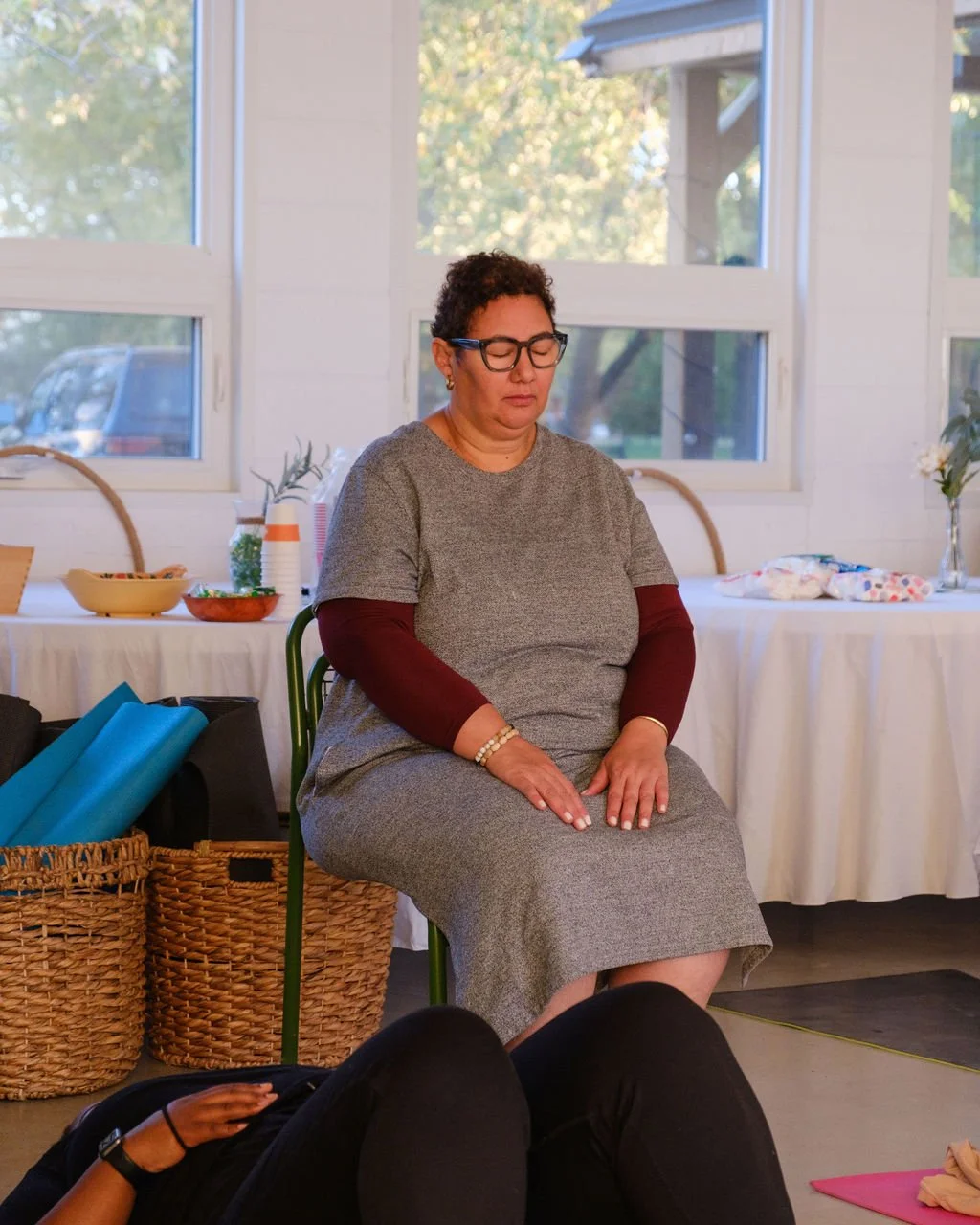 A woman meditating with closed eyes and hands resting on her lap in a room with large windows, a table with snacks and drinks, and baskets with yoga mats.