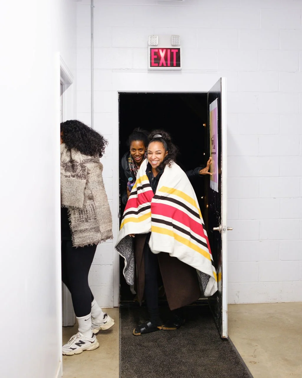 Two women exiting through a door, smiling; one woman has a colorful striped blanket draped over her shoulders; another woman with curly hair on the left side walking in the opposite direction; maximum contrast Exit sign above the door; white wall and