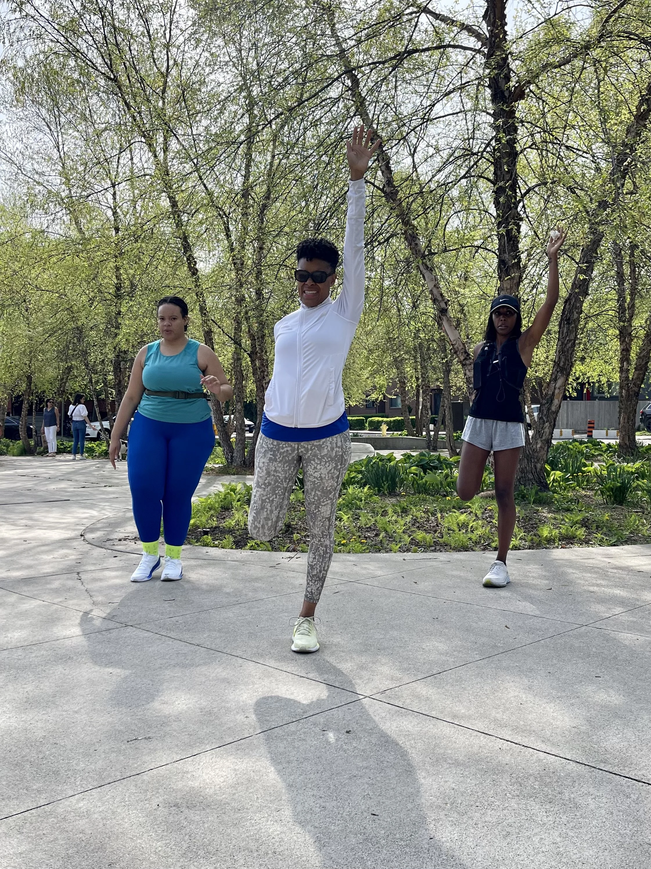 Three women exercising outdoors on a sunny day in a park with trees and green foliage.