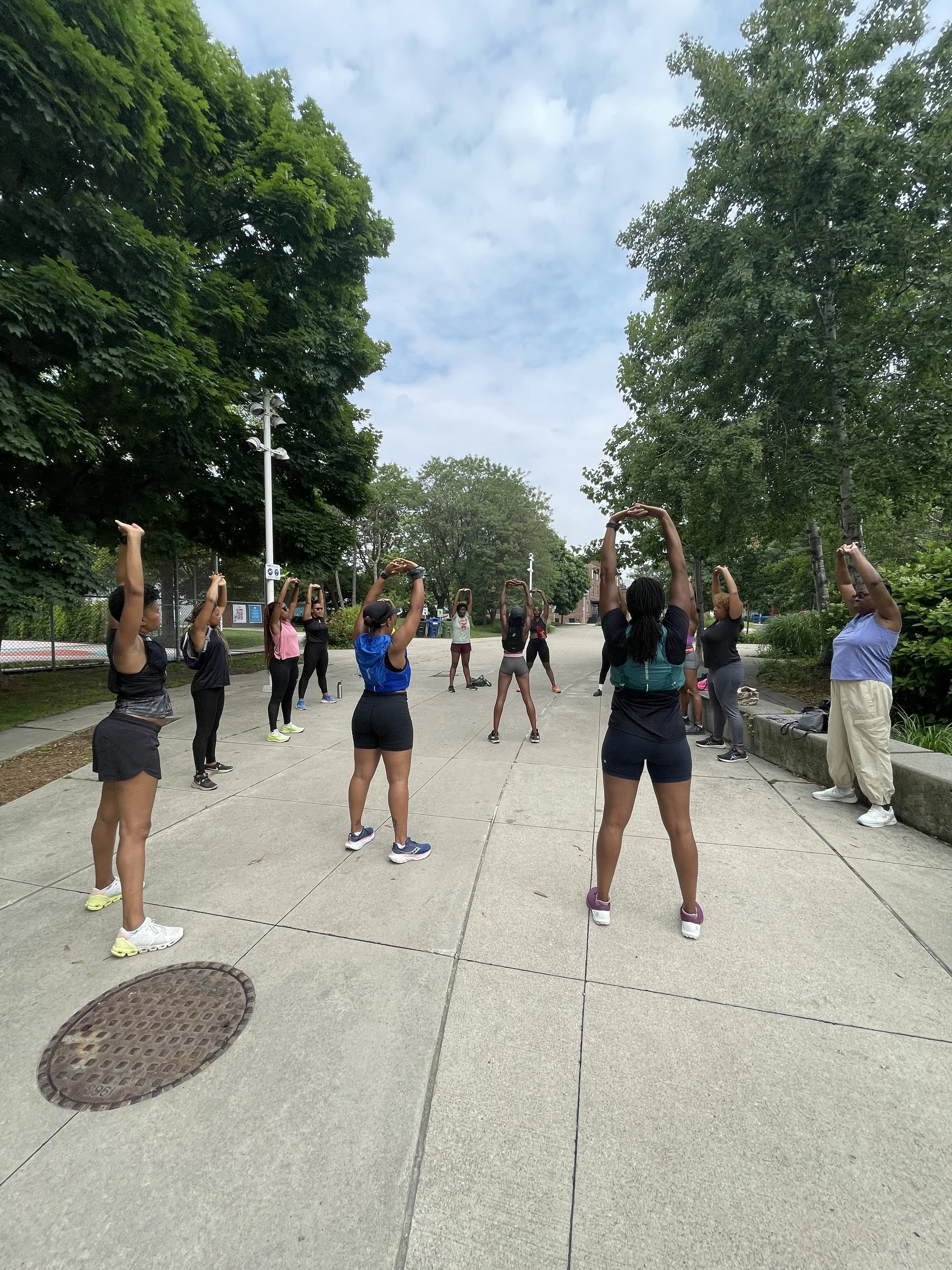 Group of women, mostly in athletic clothing, participating in an outdoor stretching or warm-up session in a park with trees and a cloudy sky.