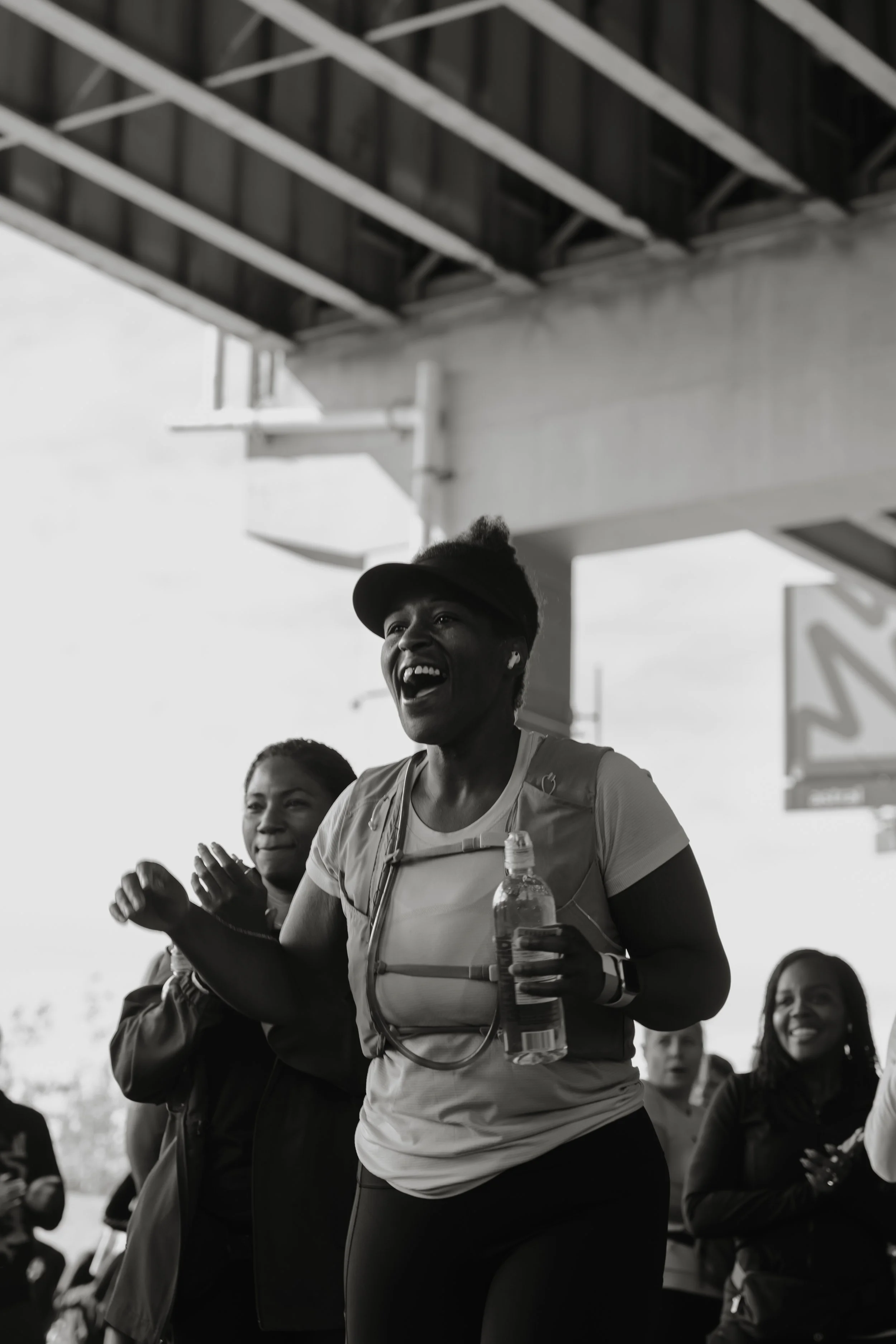 A group of women, with one smiling and holding a water bottle, appears to be celebrating or running in an outdoor setting.