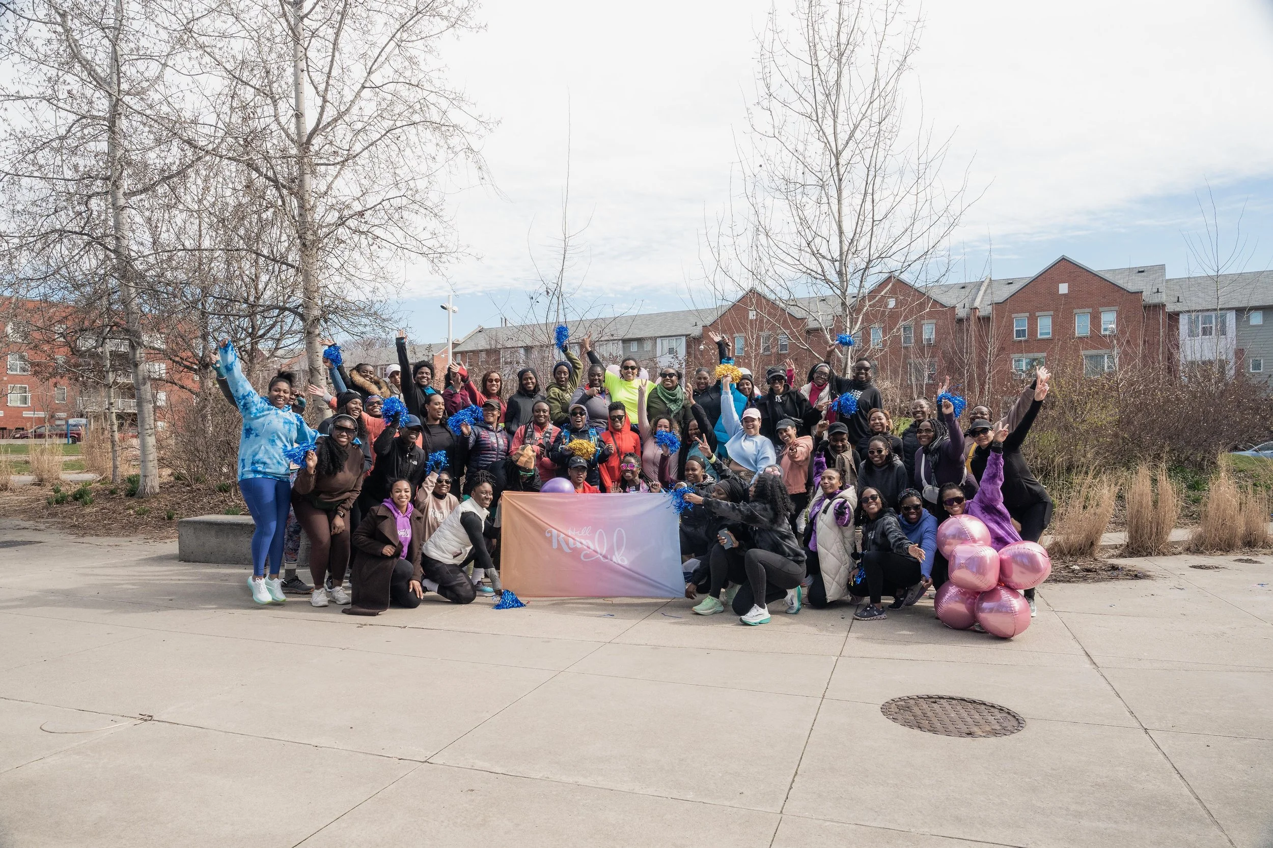 A large group of diverse people gathered outdoors on a sidewalk, holding blue and gold pom-poms, with some raising their hands. There are pink balloons on the right, and a banner in front that says 'Kanel.' The background features leafless trees and 