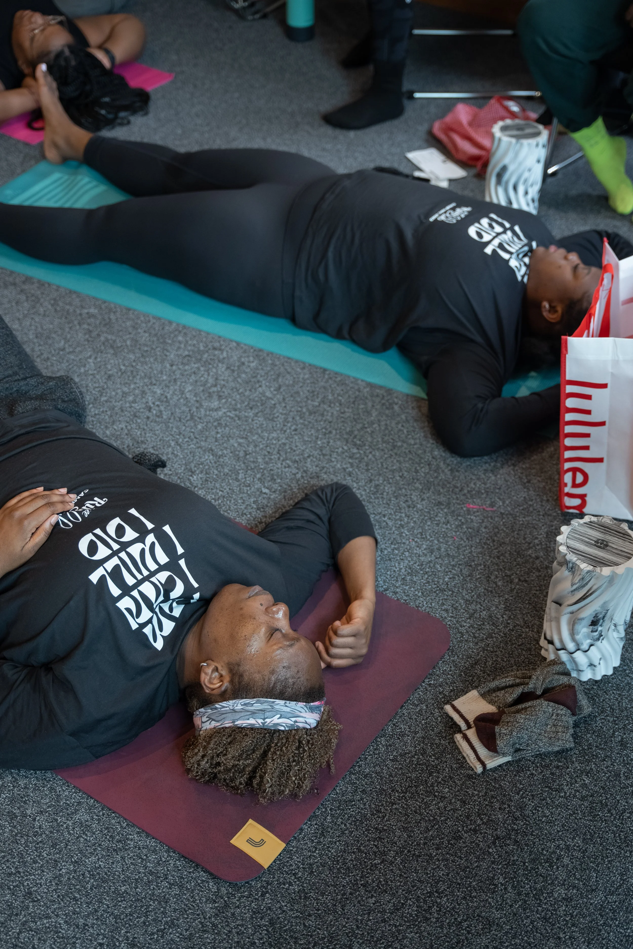 Two women lying on yoga mats indoors, engaged in a relaxation or meditation session, wearing black shirts with white text, and surrounded by personal belongings.