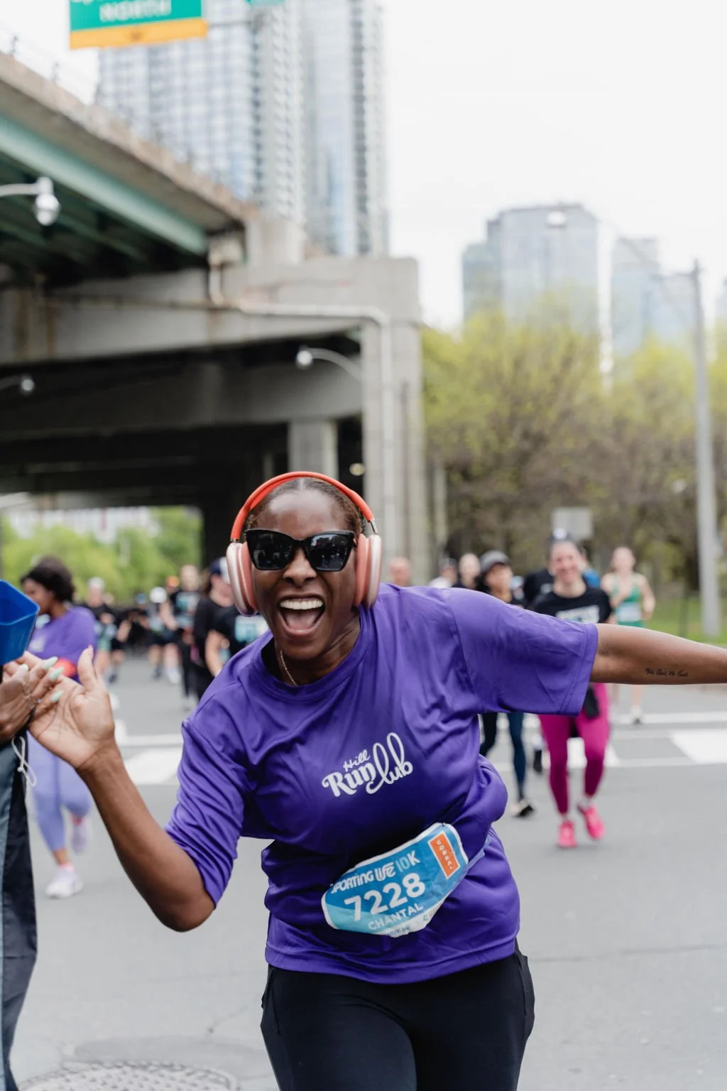 A woman with dark skin, wearing sunglasses, headphones, a purple t-shirt with white text, and a race bib number 7228, is smiling and running in a marathon or race event, with other participants and city buildings in the background.