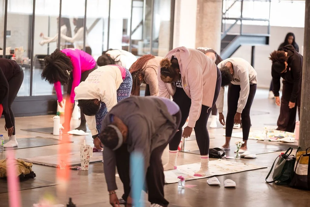Group of people participating in a yoga or meditation class, bending forward in a spacious indoor setting with large windows.