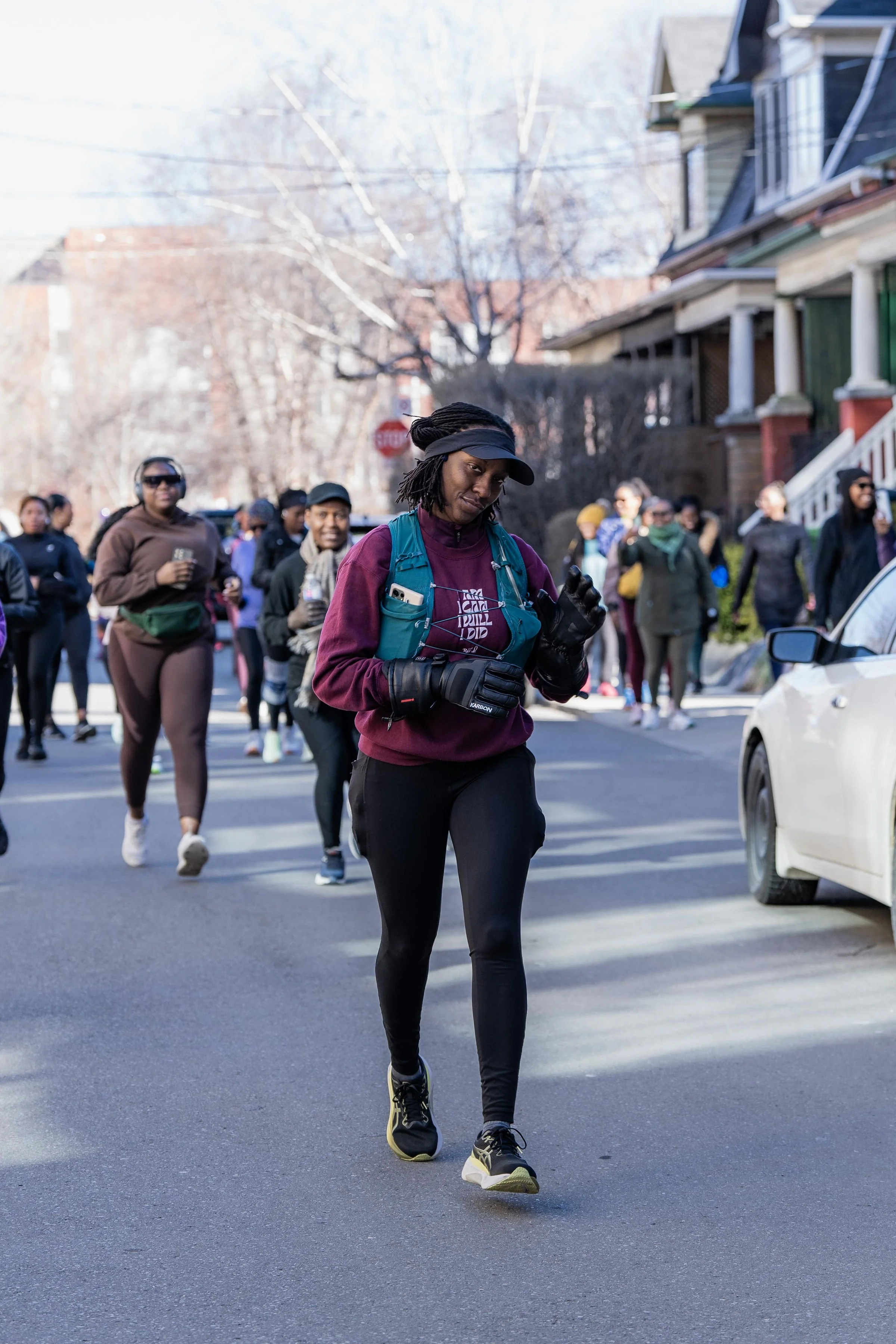 A woman in athletic clothing and black gloves running in a group on a city street during fall or winter, with houses and bare trees in the background.