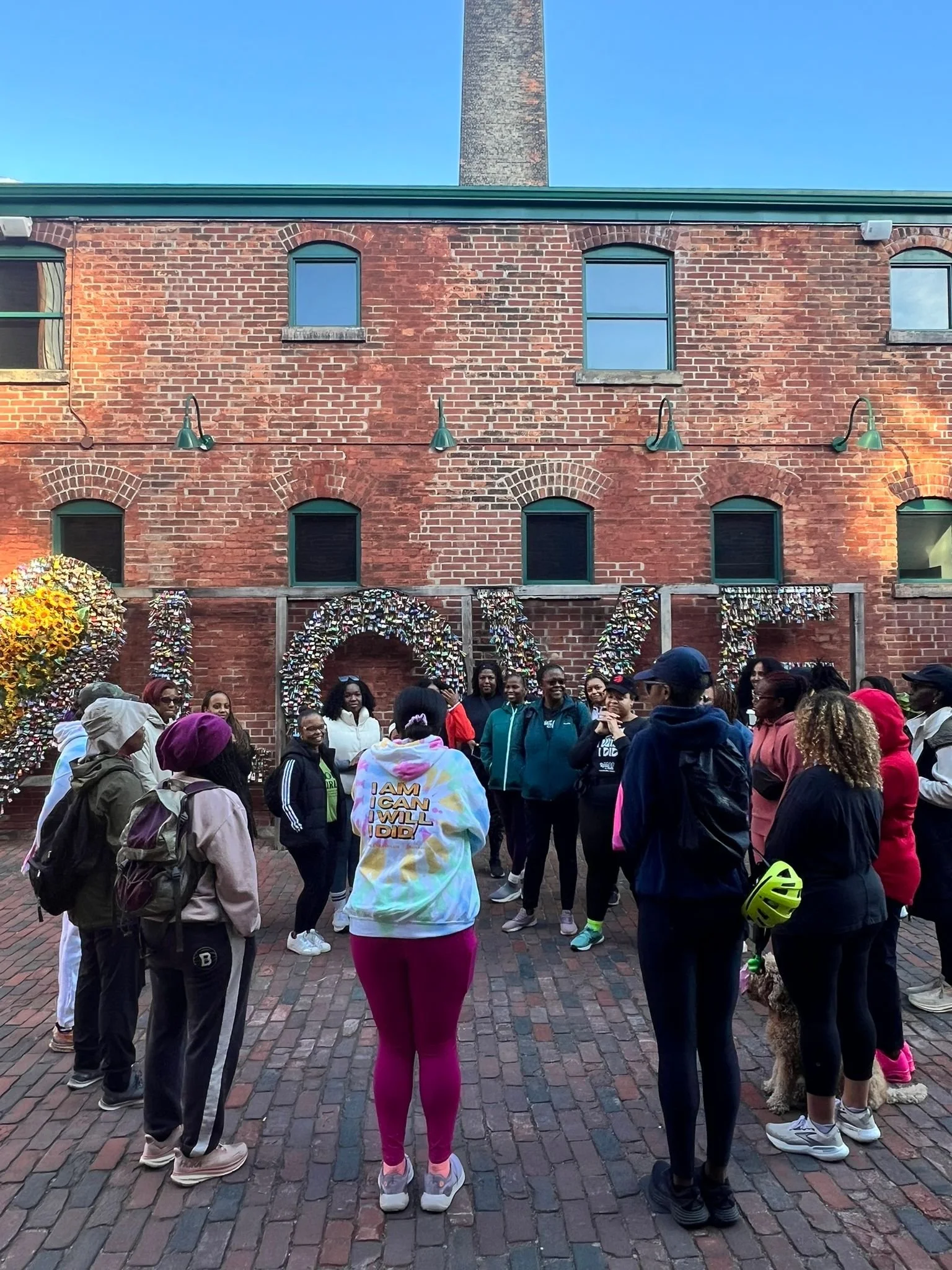 Group of diverse people gathered outside a brick building with large decorative letters spelling 'LOVE' made of flowers and objects, possibly for a community event or assembly.
