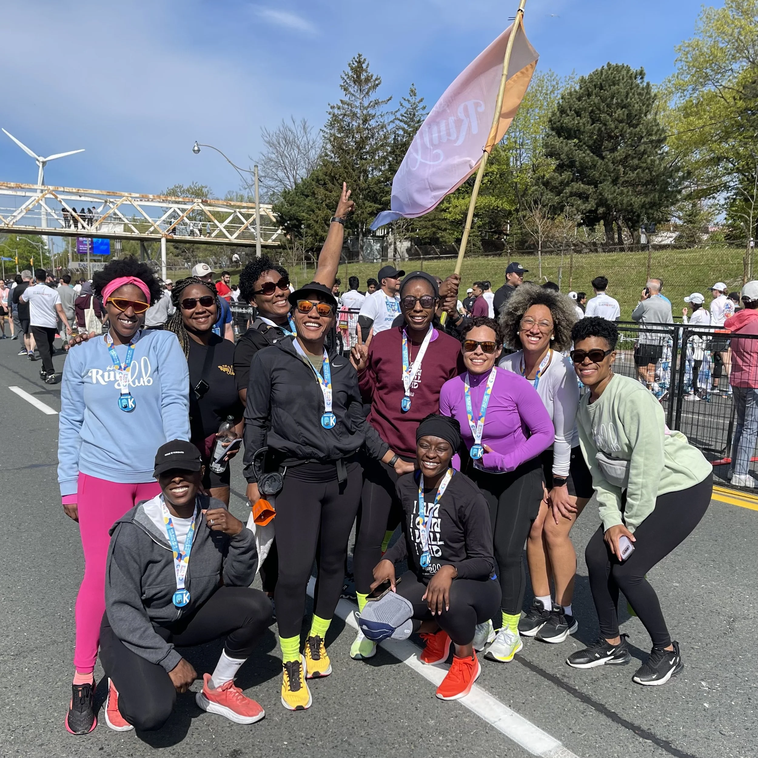 A group of women with medals around their necks, smiling and posing for a photo at a race event. One woman is holding a pink flag, and they are all dressed in athletic clothing, with a background of other participants and trees.