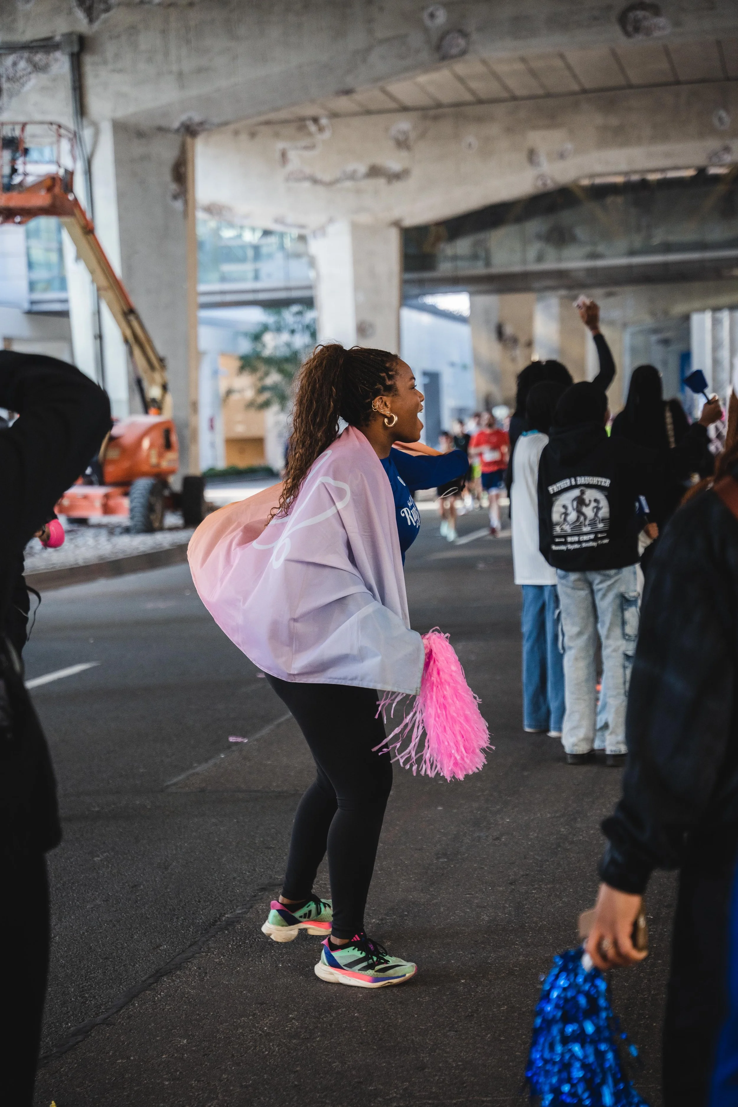 A woman cheerleader with a pink pom-pom on her wrist, standing on a street under a bridge, cheering with a group of people during a parade or event.