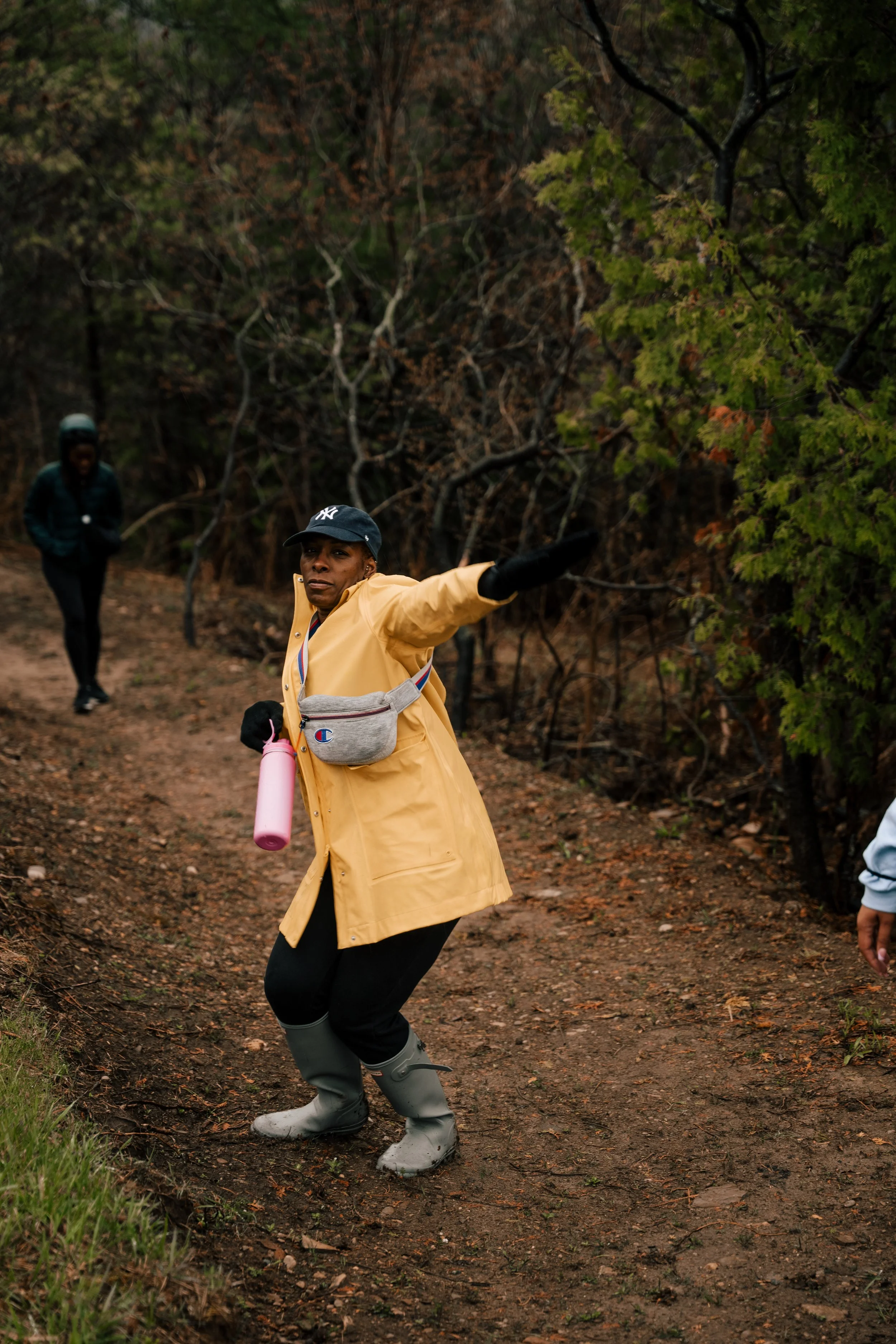Woman in yellow raincoat and rain boots directing while on a forest trail, with two other people in the background.