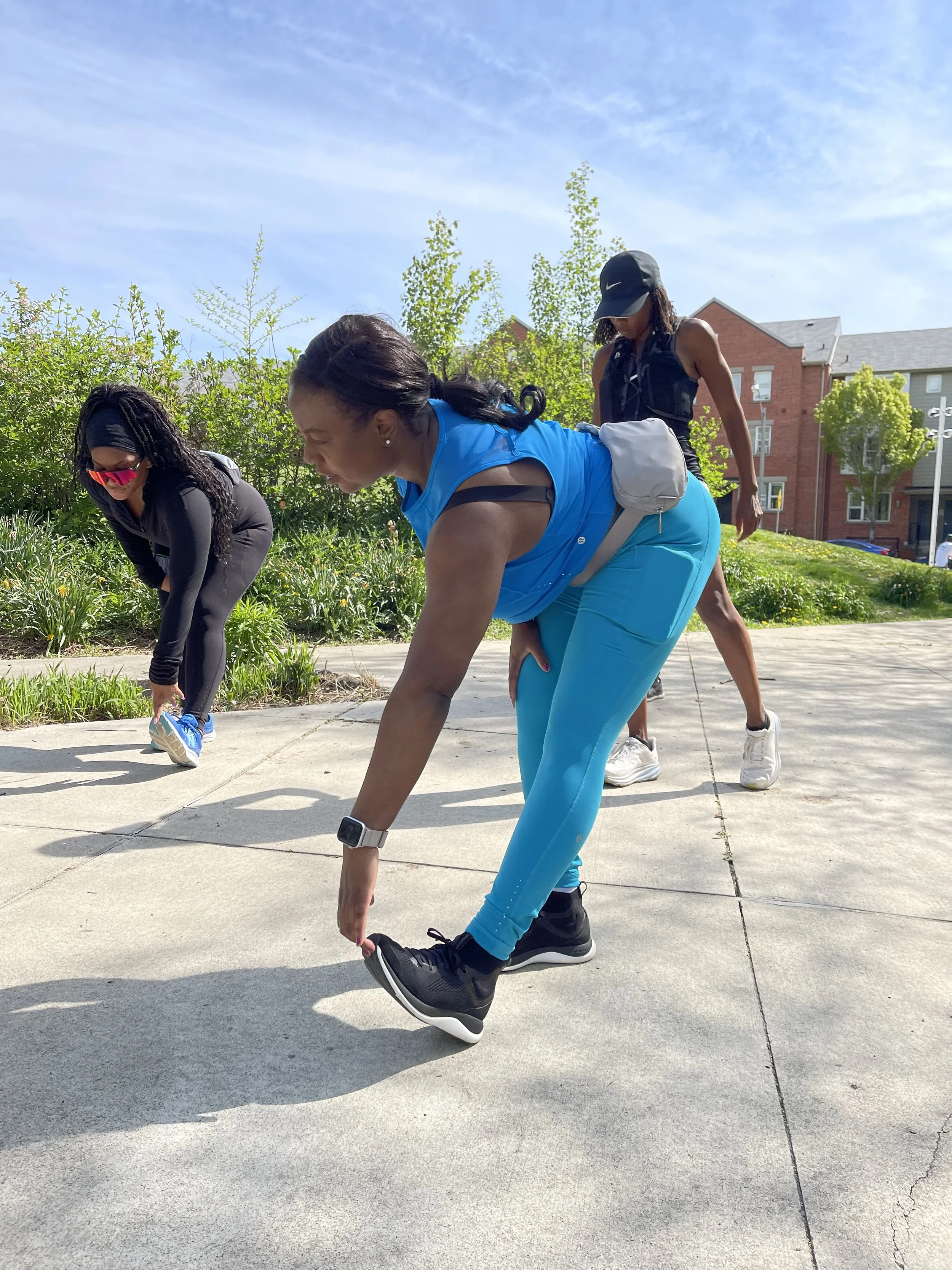 Three women stretching outdoors on a sidewalk during a sunny day. They are wearing athletic clothing and sneakers.