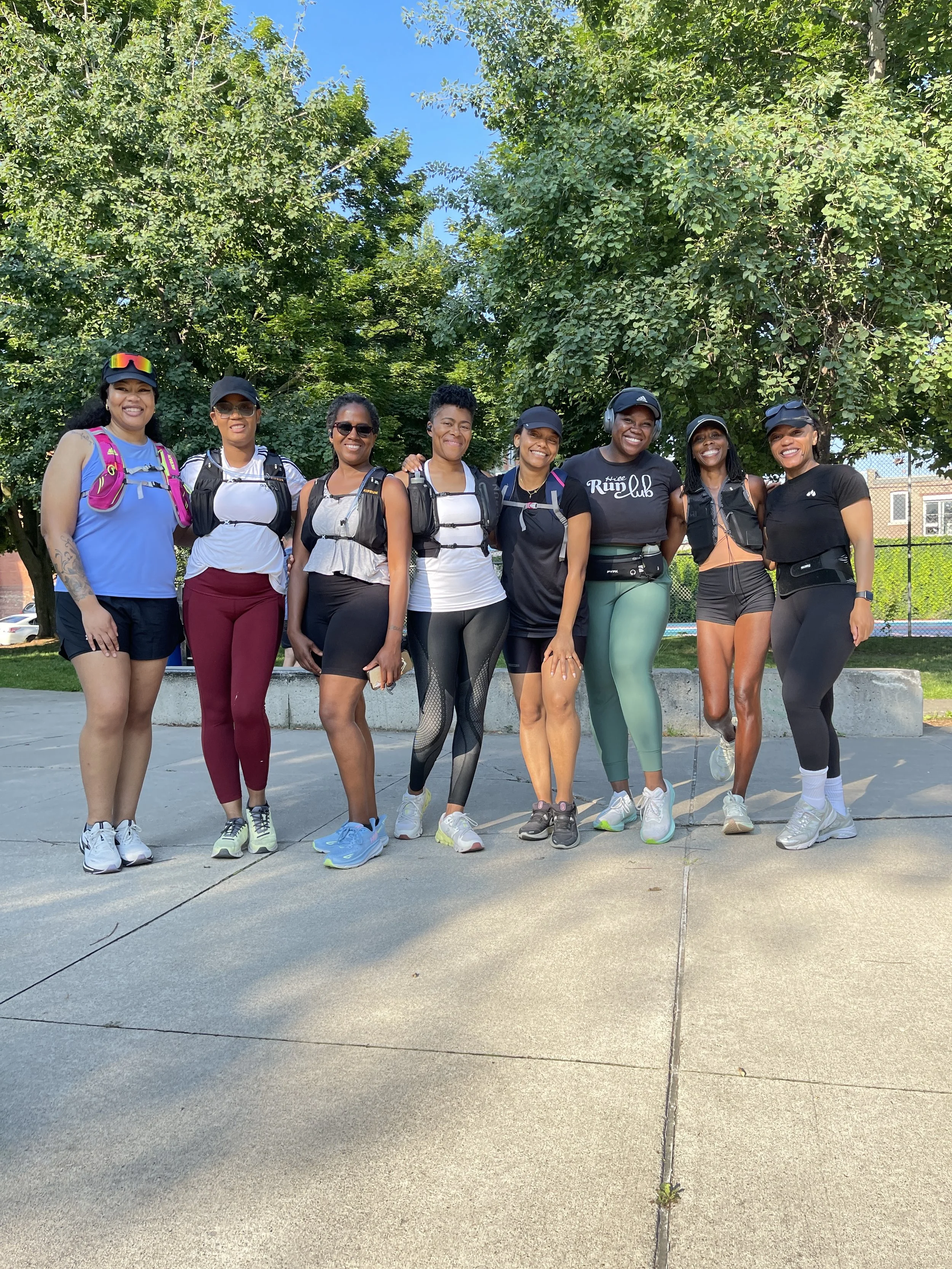 A group of nine diverse women standing outdoors in athletic clothing and gear, smiling with trees in the background.