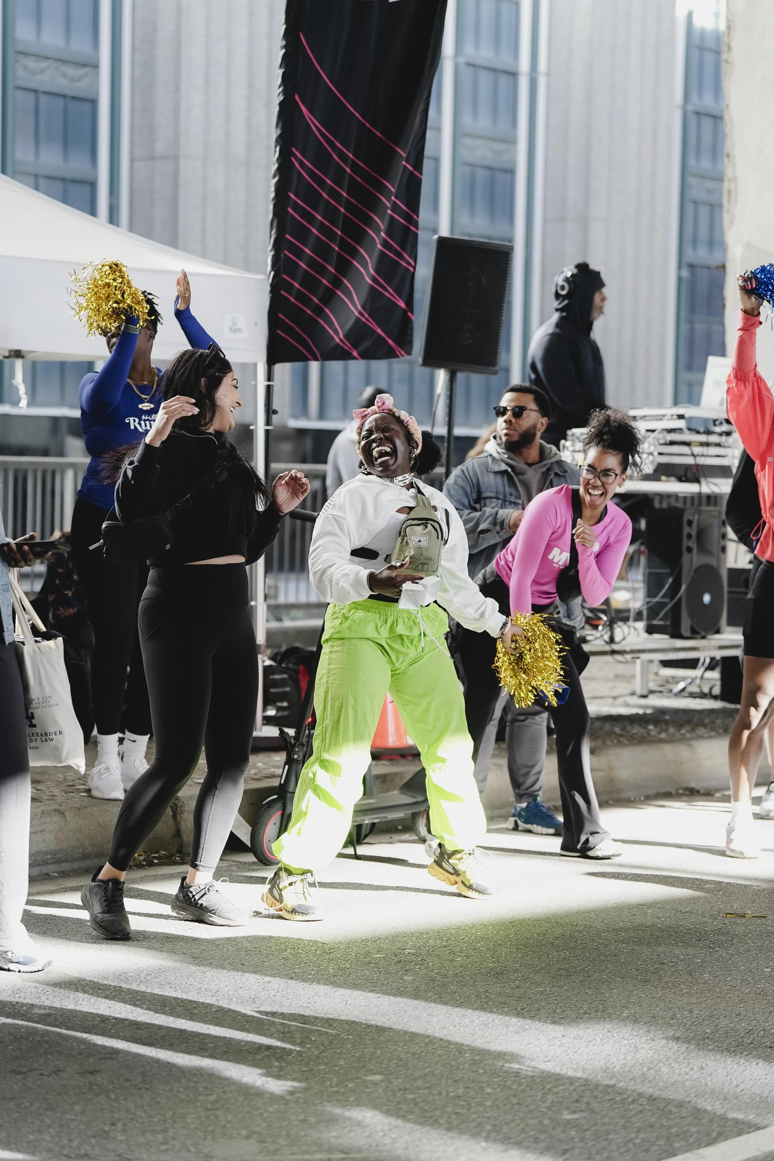Group of people celebrating on a city street, some holding gold pom-poms, with a DJ setup and tall city buildings in the background.