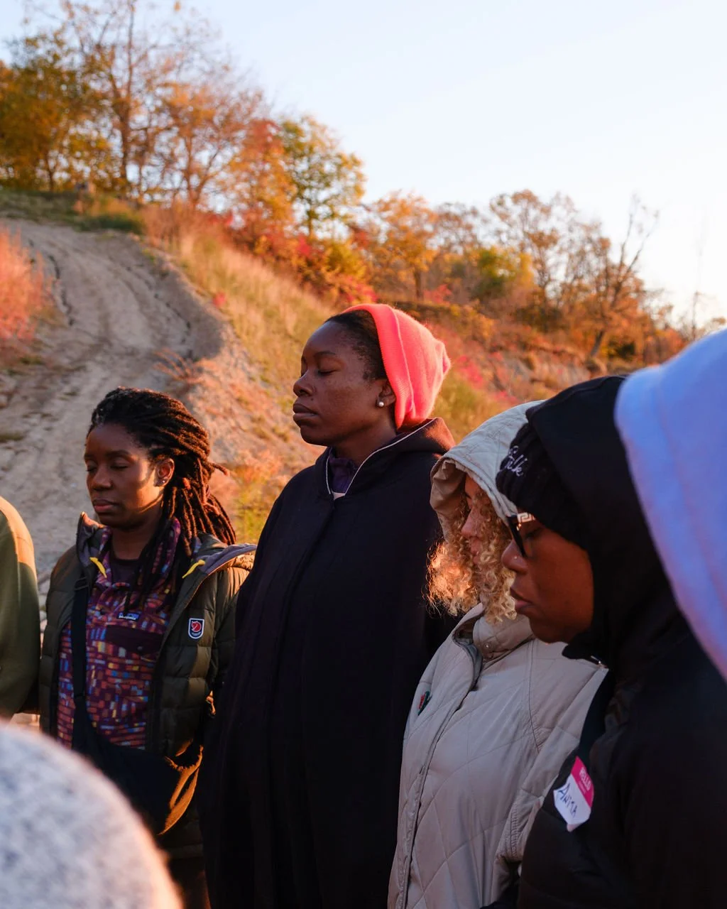 Group of diverse women with closed eyes outdoors during sunset, standing in a prayer or meditation circle.