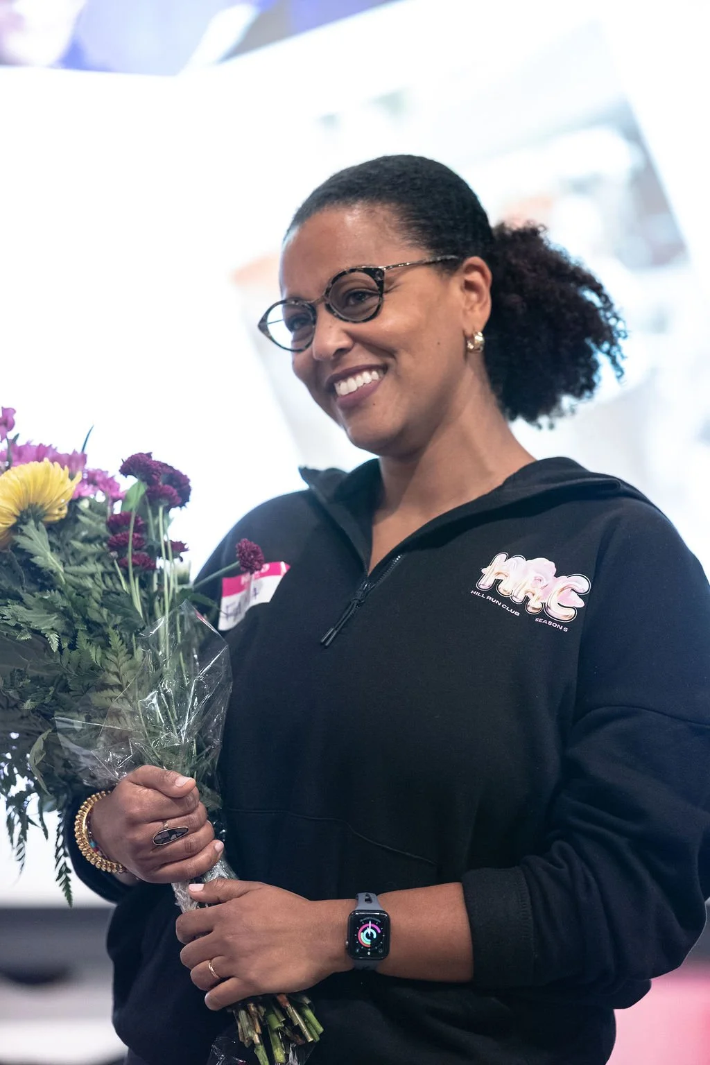 A woman smiling, holding a bouquet of colorful flowers, wearing glasses, a black hoodie with a logo, a smartwatch, and a ring, indoors with a bright background.