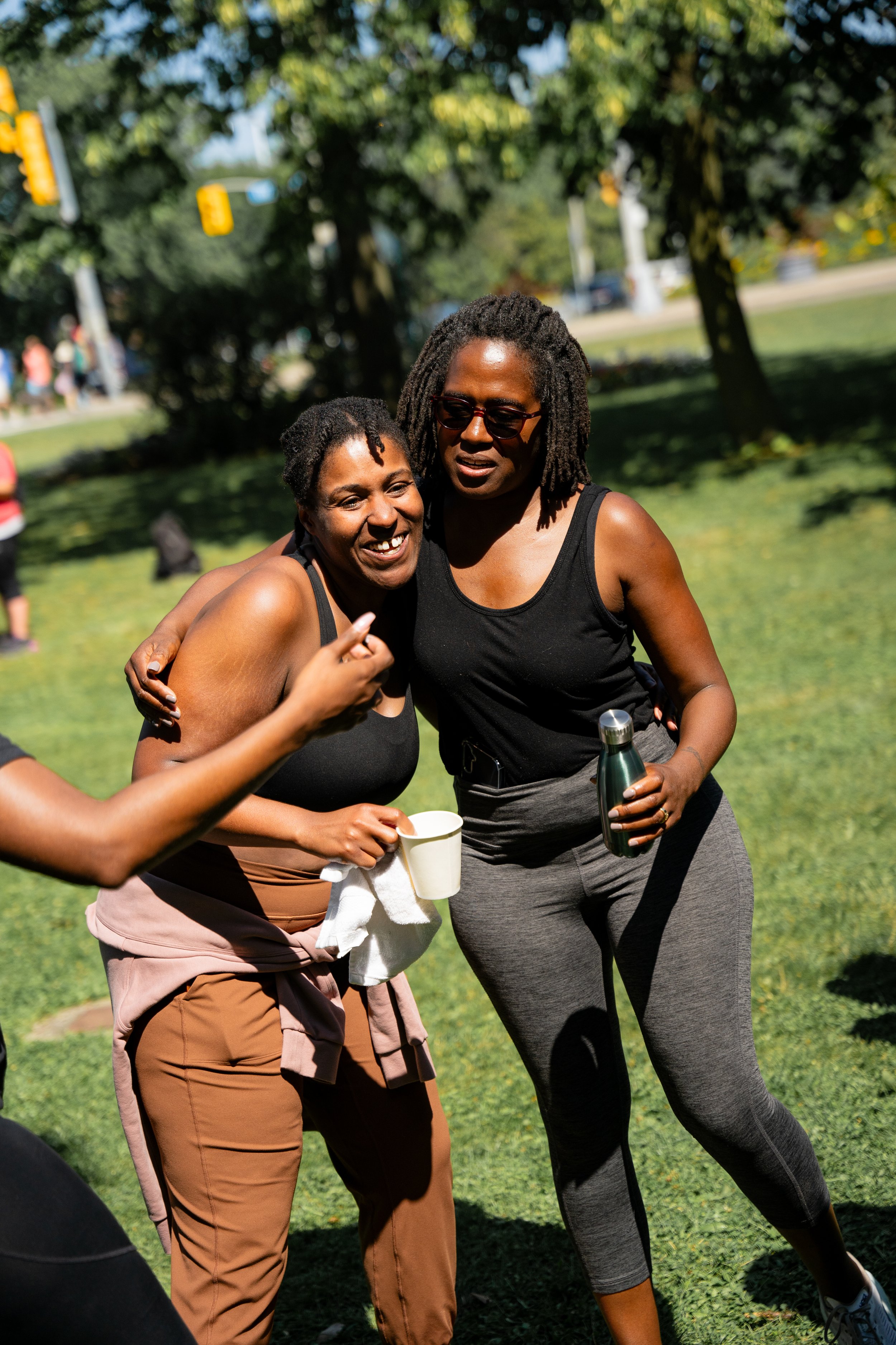 Two women wearing workout clothes hugging and smiling outdoors in a park on a sunny day.