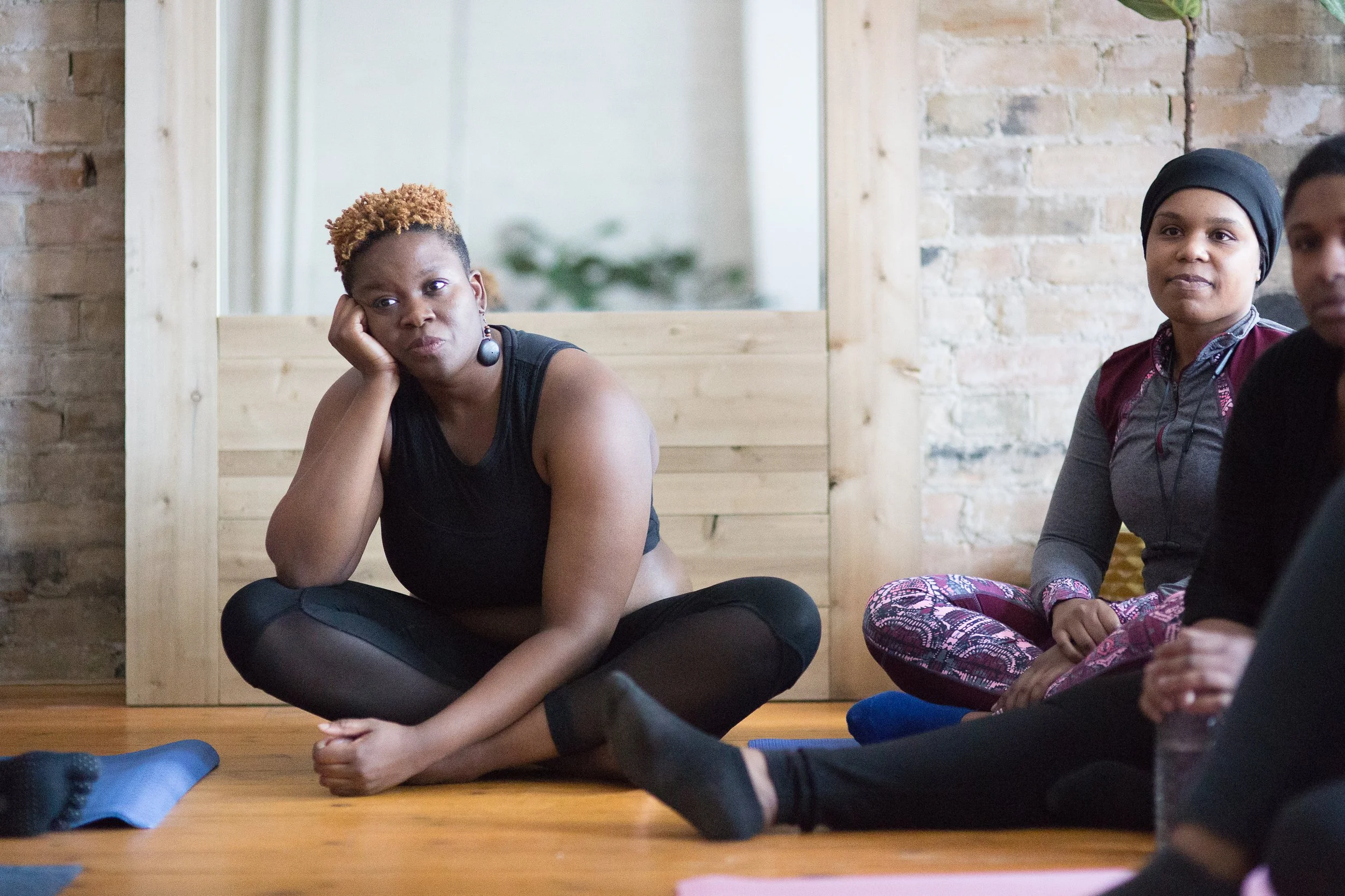 Women sitting on the floor during a yoga or meditation class in a room with wooden flooring and brick walls.