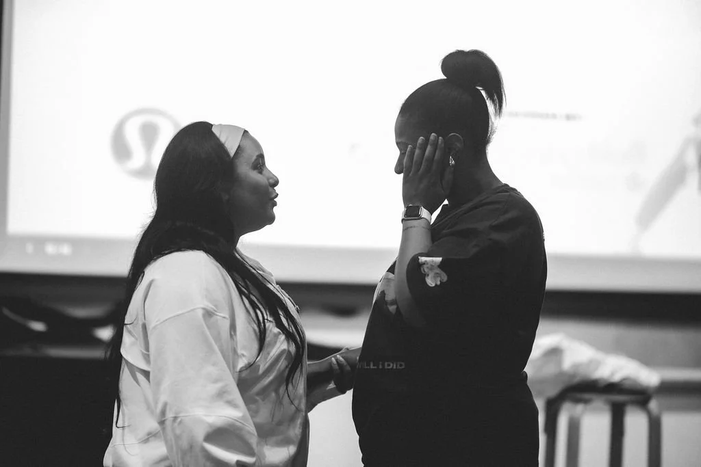 Two women standing face-to-face in a room, one with long dark hair and a headband, the other with her hair in a bun, appears emotional and comforting each other.