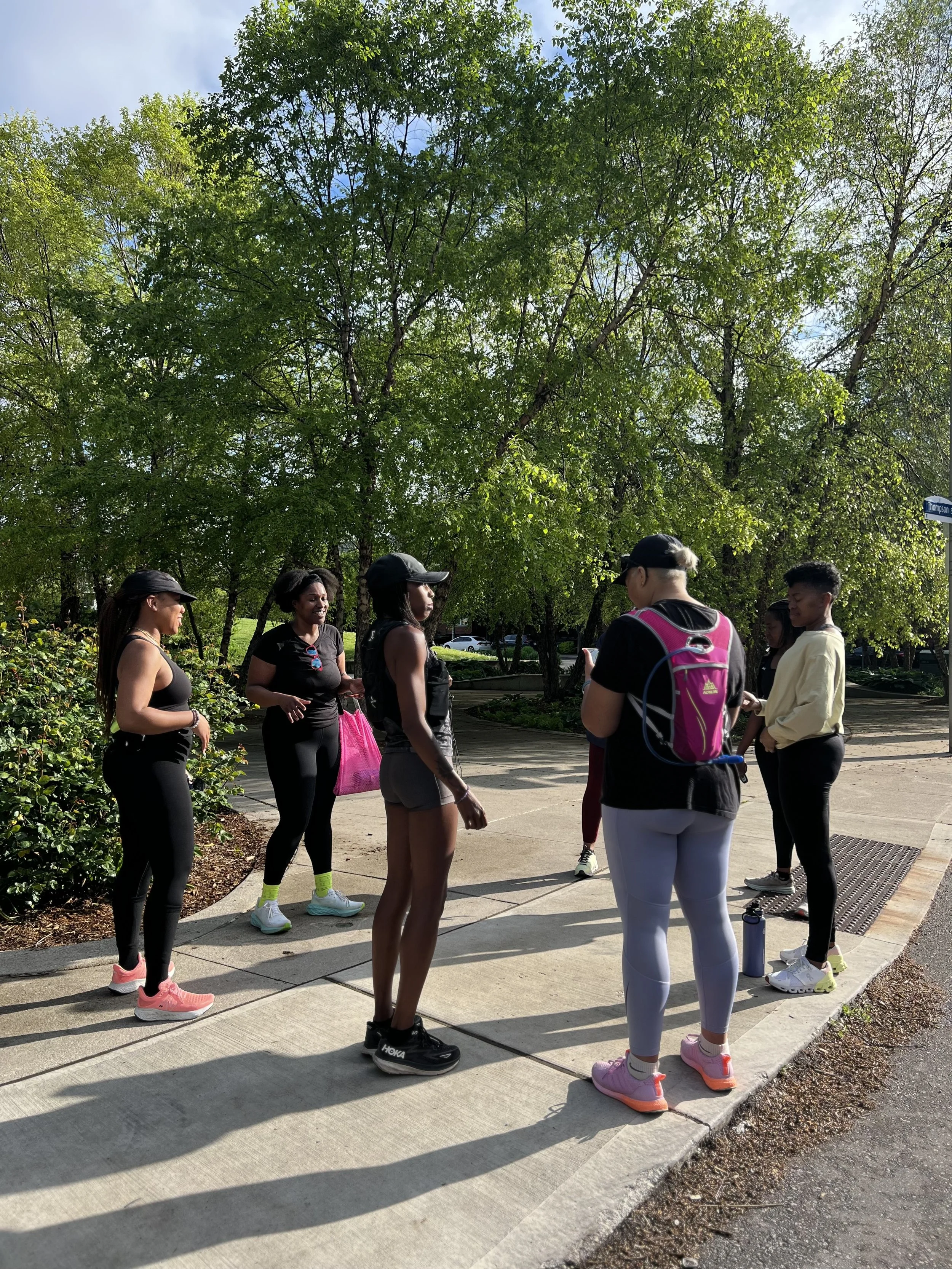 Group of six women and one man in athletic clothing standing on a sidewalk in a park, engaging in conversation with lush green trees and a street sign in the background.