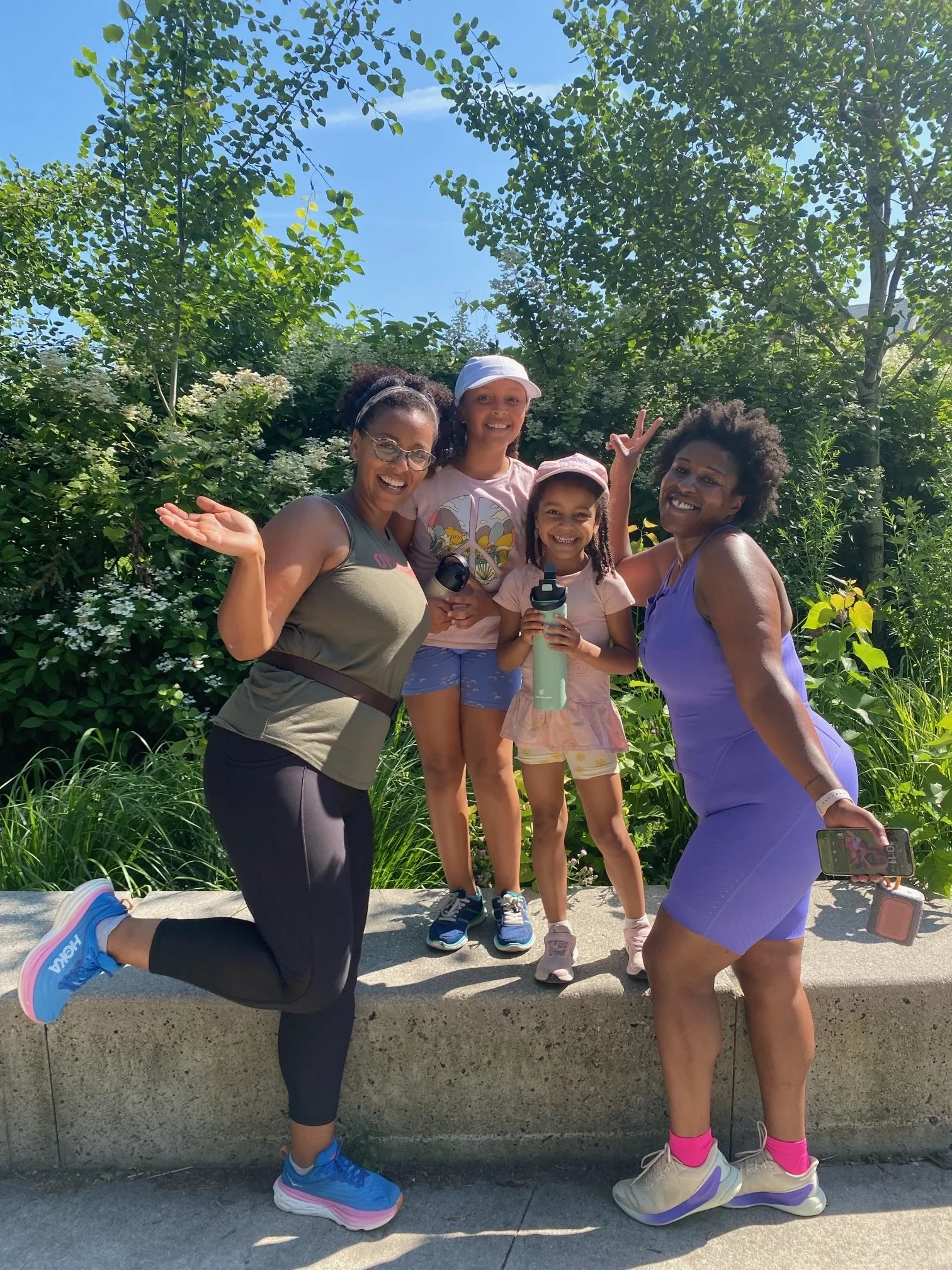 Four women and two young girls outdoors on a sunny day, standing on a concrete ledge with green trees and bushes behind them. The women are smiling and posing happily, with one woman wearing a purple dress and holding a phone, the other wearing athle
