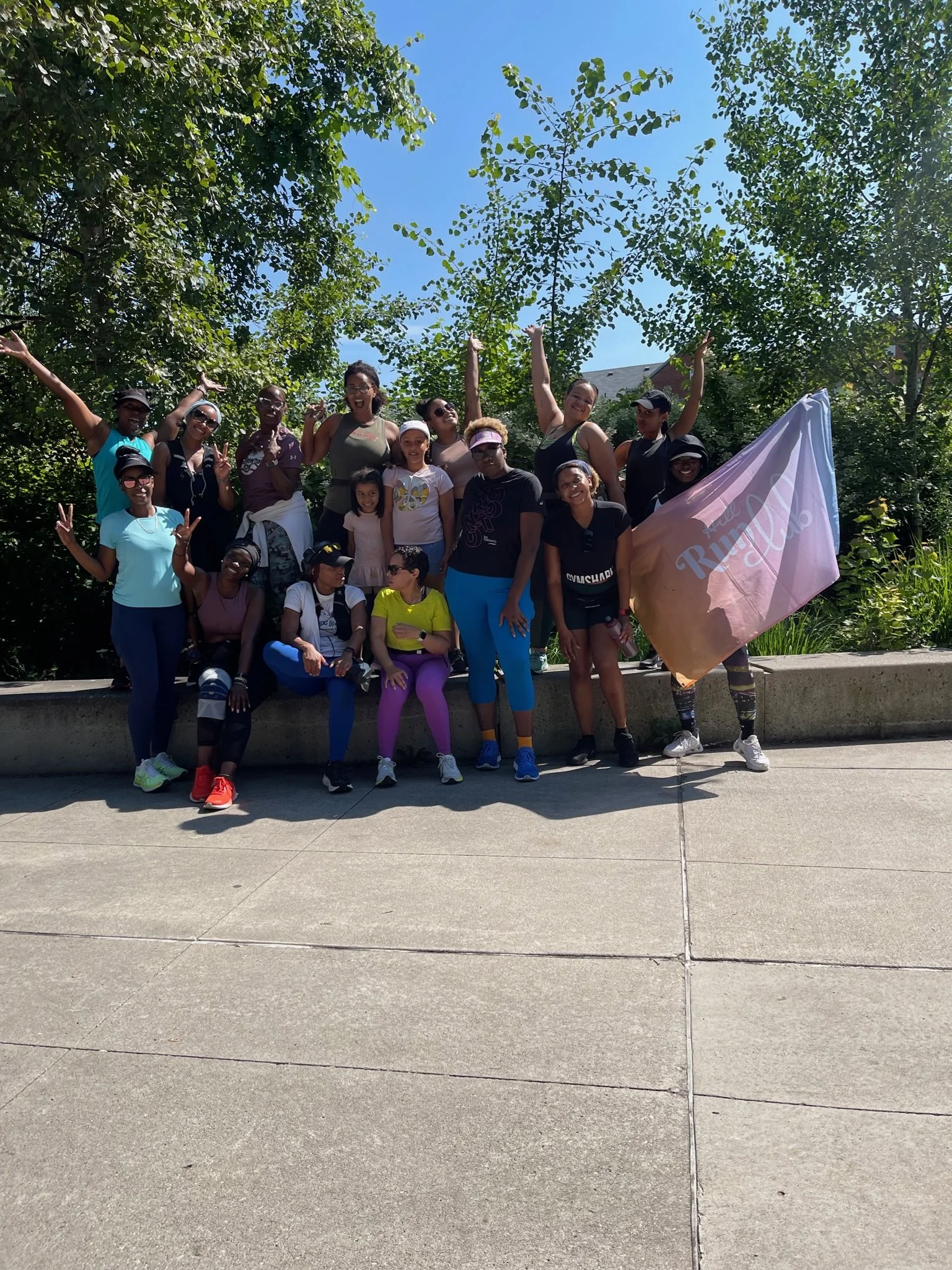Group of people posing outdoors on a sidewalk, surrounded by trees, with one person holding a pink flag that reads 'Run for the Roses' under a clear blue sky.