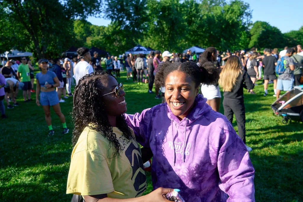 Two women smiling and hugging outdoors at a crowded event with trees and tents in background.