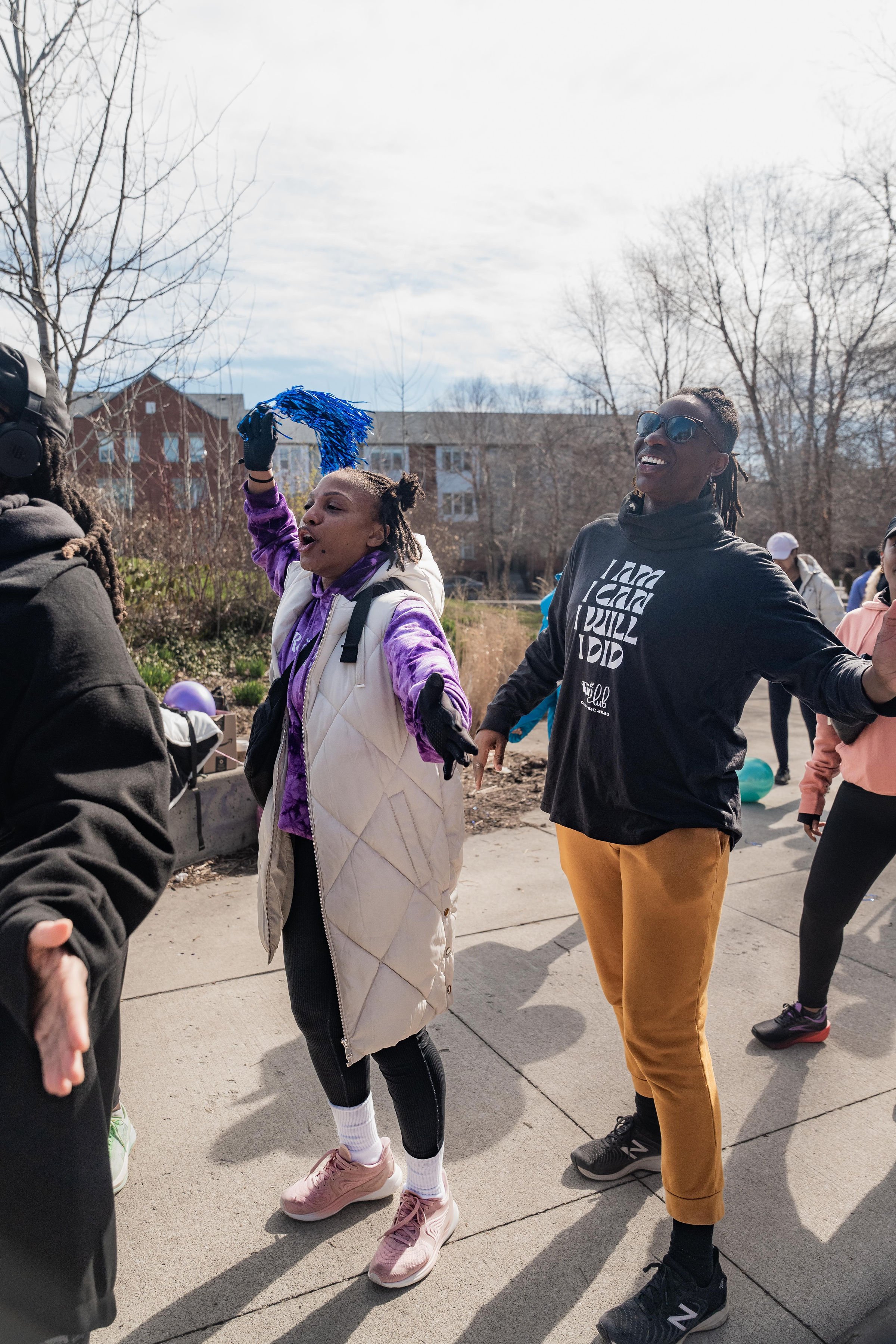 People participating in an outdoor event, dancing with one woman holding blue pom-poms, on a cloudy day with bare trees and buildings in the background.