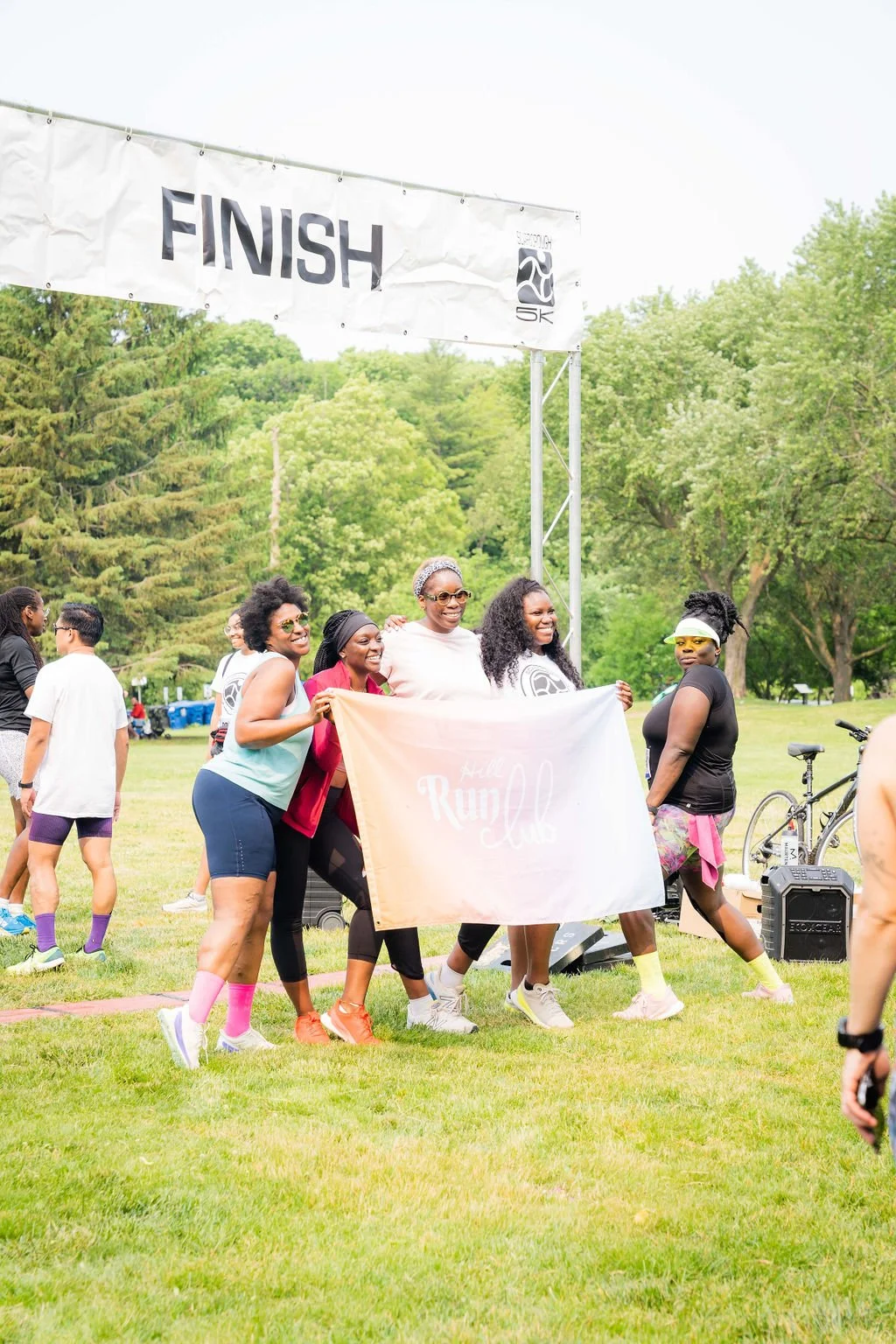 Group of women at a park holding a banner that says 'Hell Run Club,' celebrating near a finish line.
