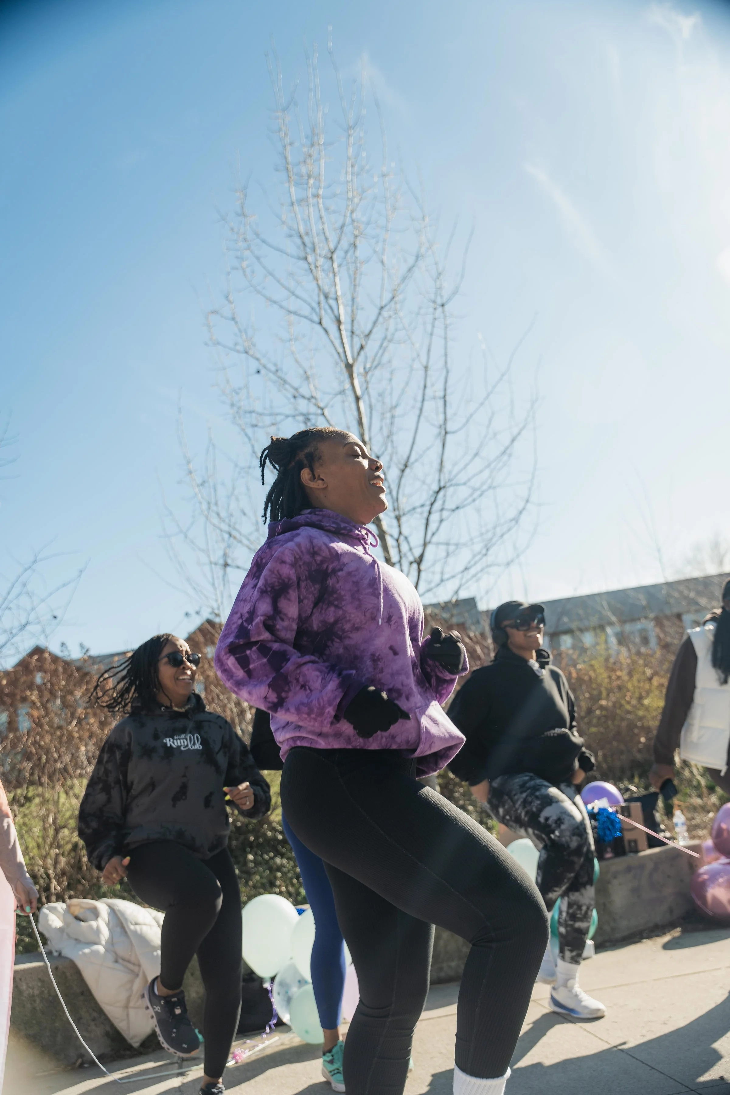 A group of women jogging outdoors on a sunny day, with balloons and trees in the background.