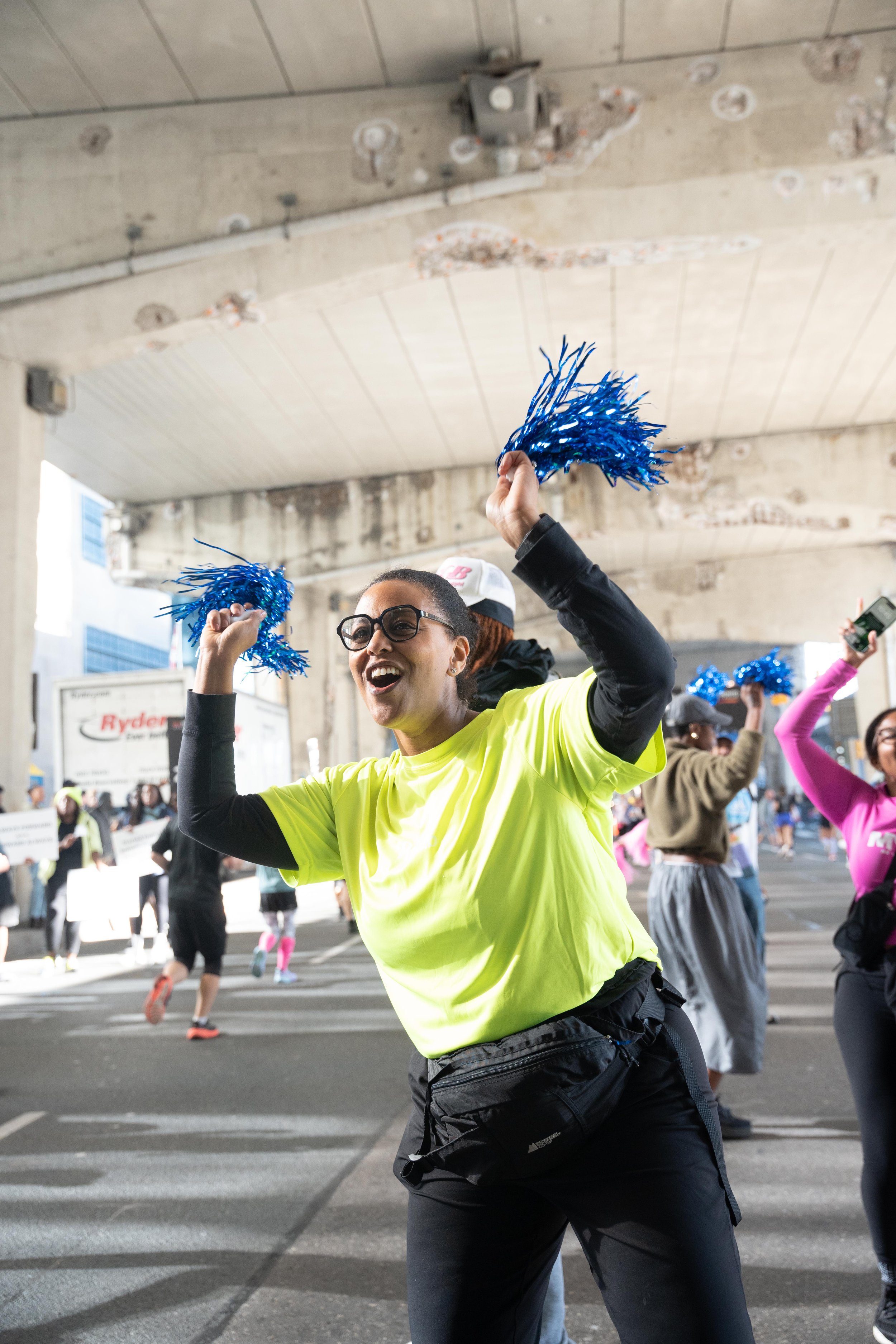A woman wearing glasses, a neon yellow shirt, and black pants celebrating at a public event under a concrete overpass, holding blue pom-poms.