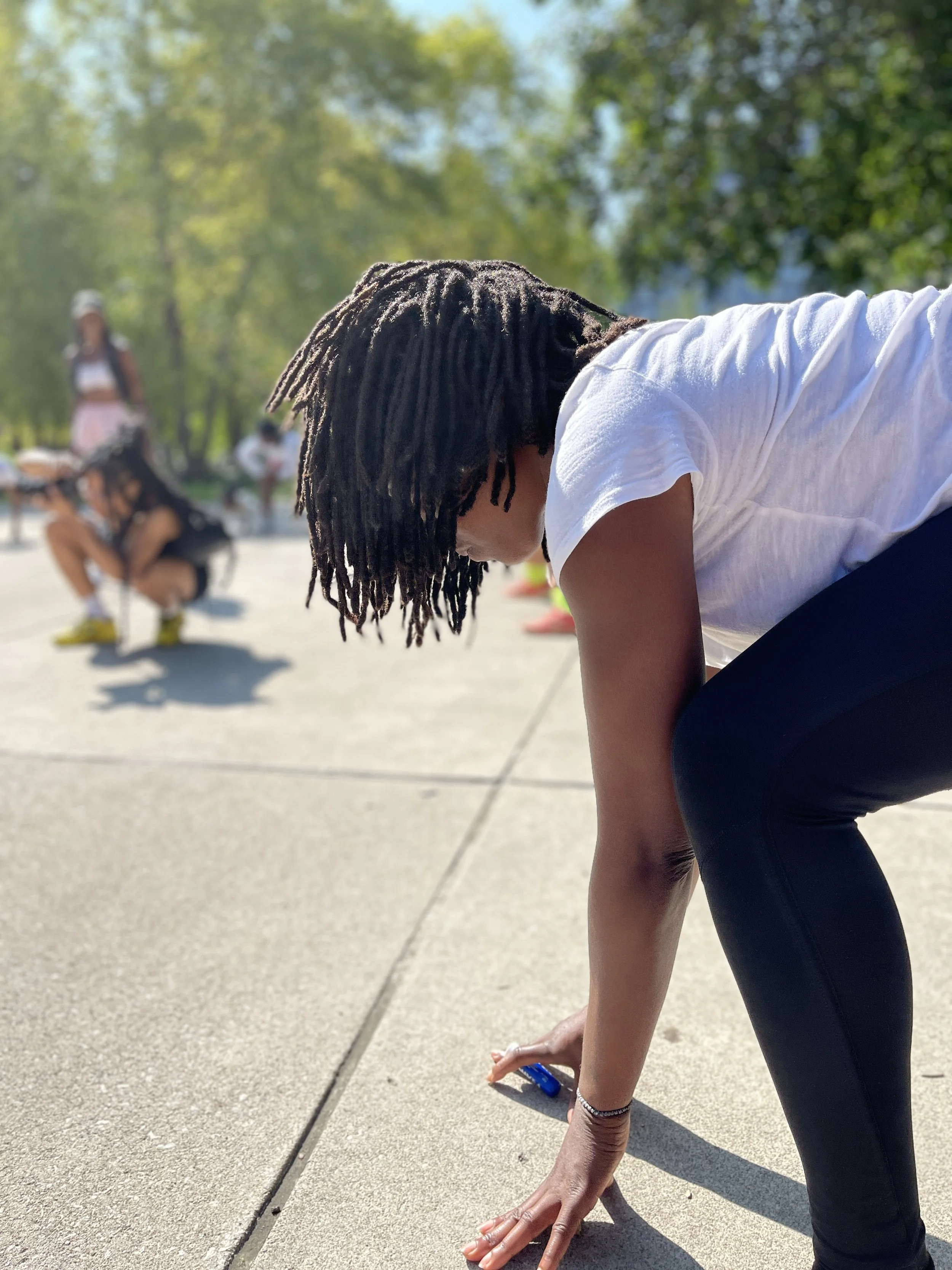 Person with dreadlocks in white T-shirt and black leggings in a squat pose outdoors on a sunny day, with trees in the background and other people visible in the distance.