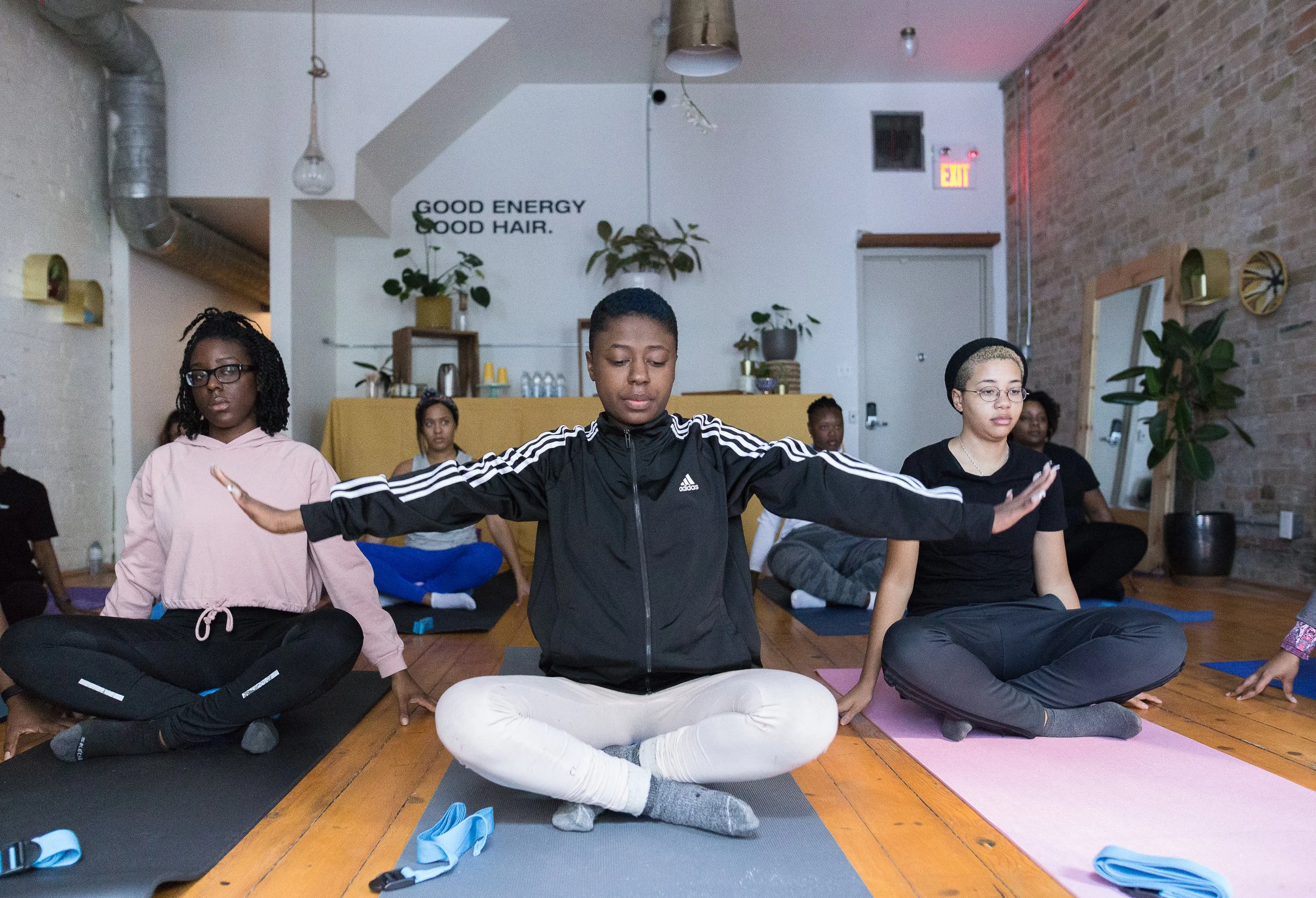 Group of women participating in a yoga class, sitting cross-legged on yoga mats with eyes closed, in a studio with exposed brick walls and potted plants.