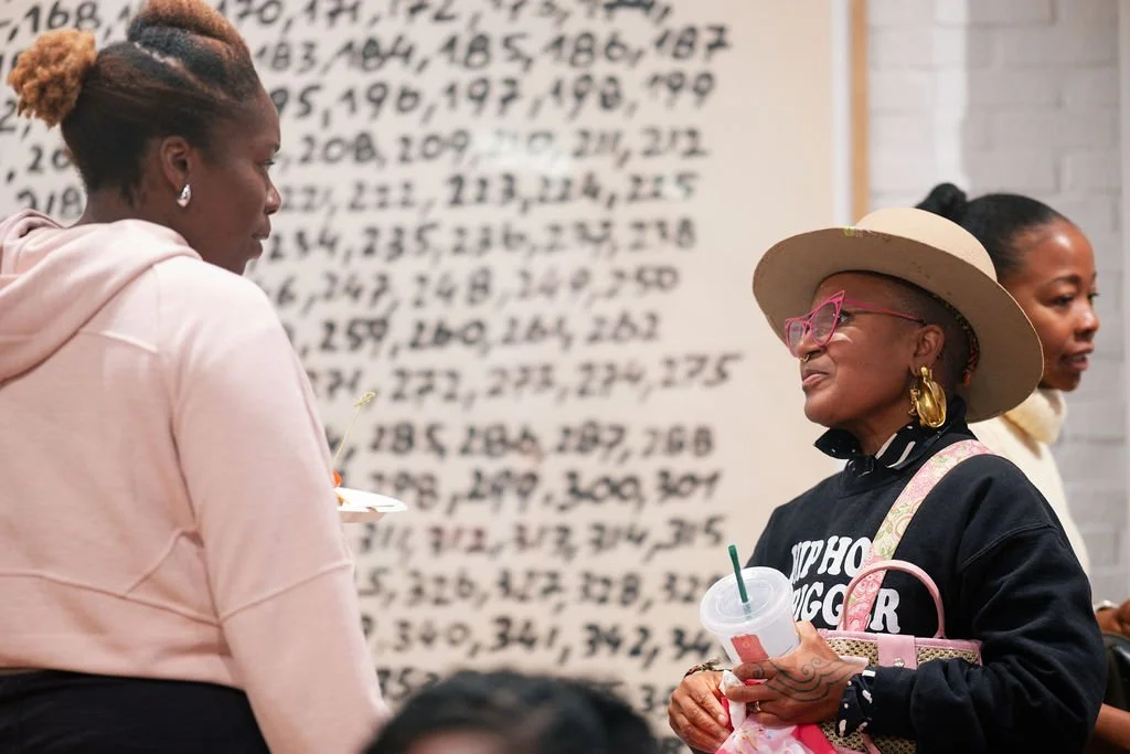 Three women standing and talking in a room with a wall filled with handwritten numbers in the background. The woman in the foreground wears a wide-brimmed hat, pink glasses, large earrings, a black sweatshirt, and carries a pink purse and a drink. Th