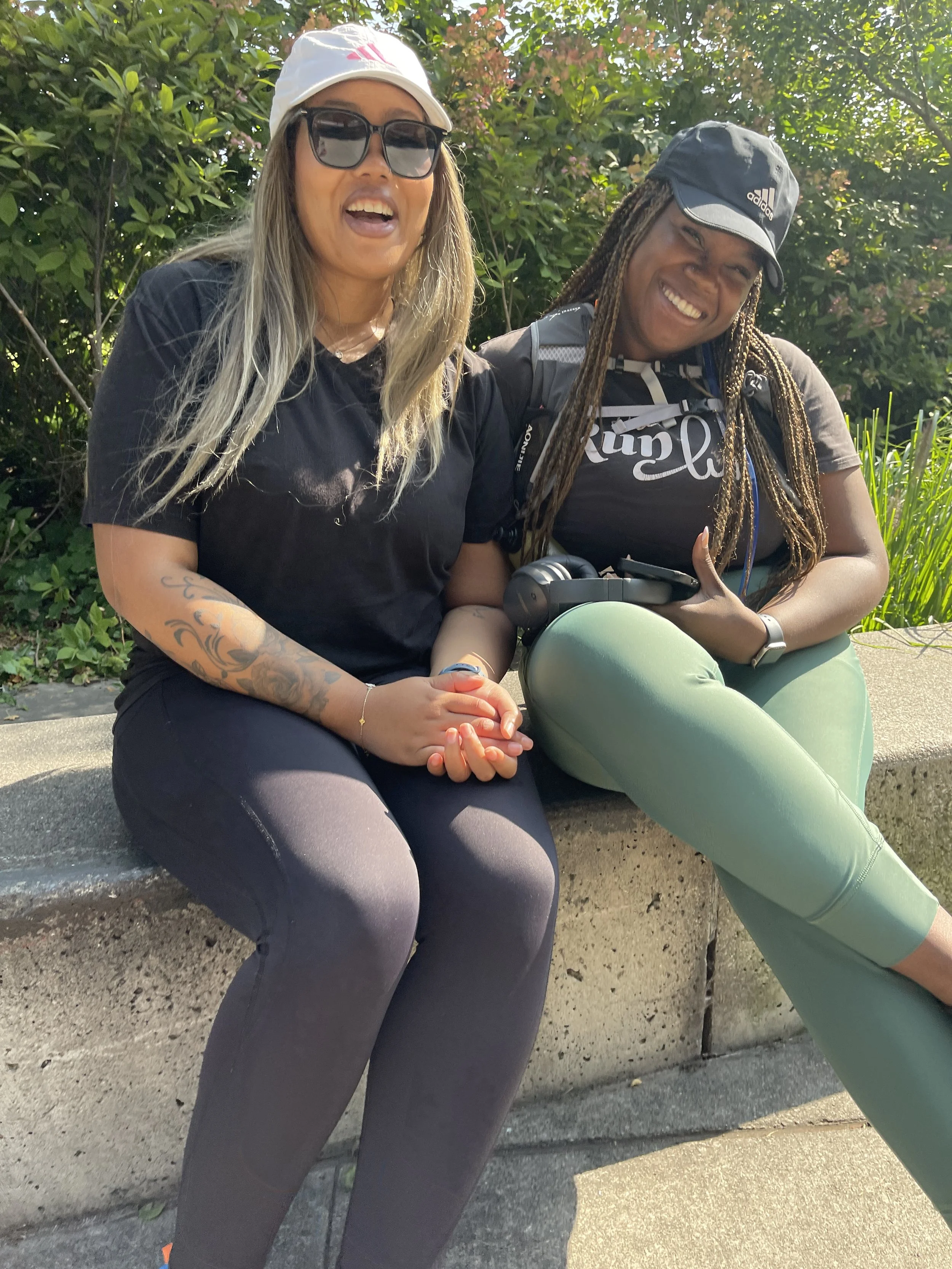 Two women sitting on a concrete bench outdoors, smiling and enjoying a sunny day, with greenery in the background.