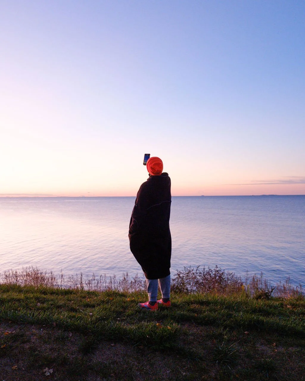 Person standing on grass near water at sunset, wearing a red beanie and black coat, taking a photo with a smartphone.