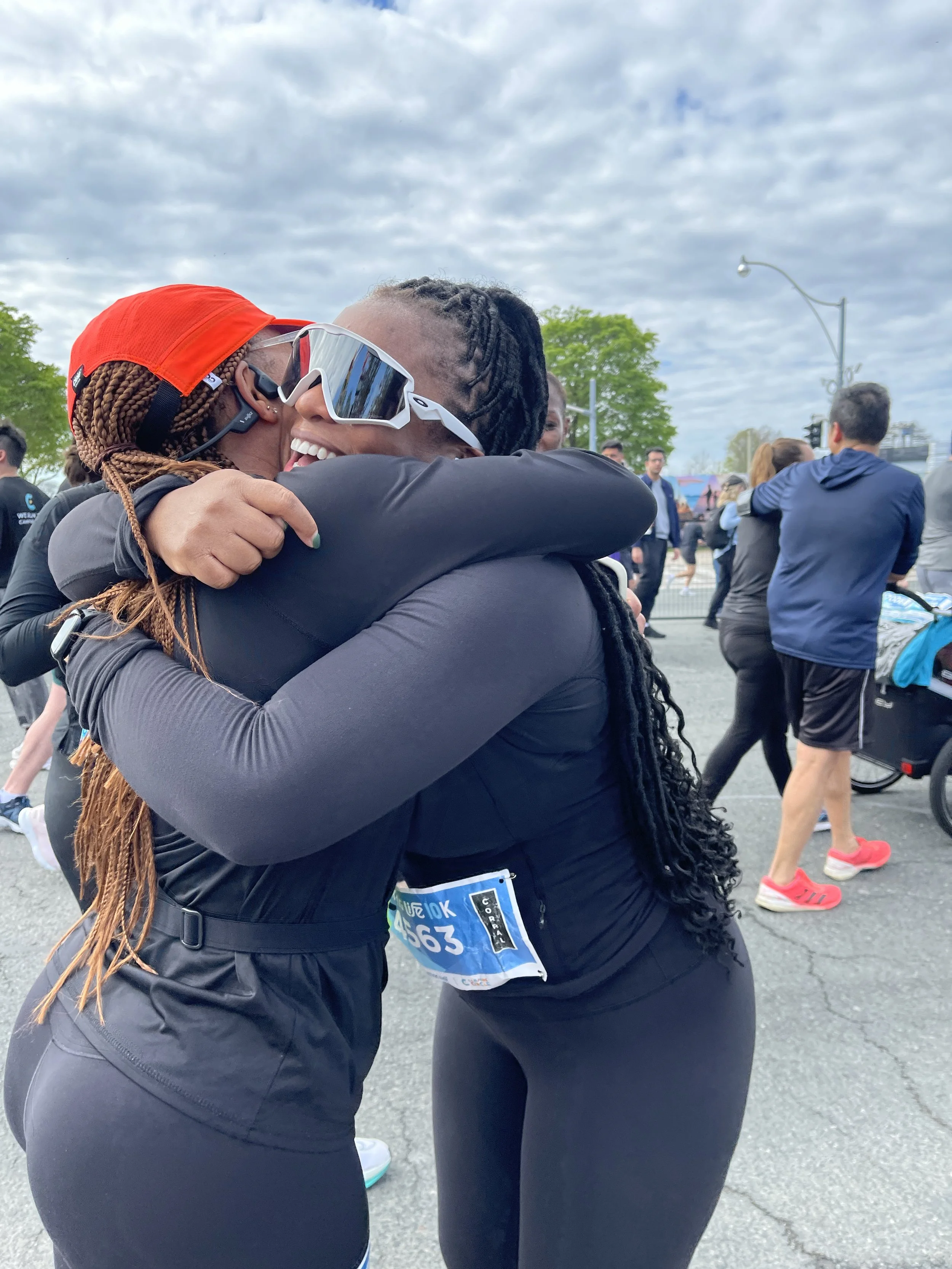 Two women in athletic gear hugging after a race, with other people in the background at an outdoor event.