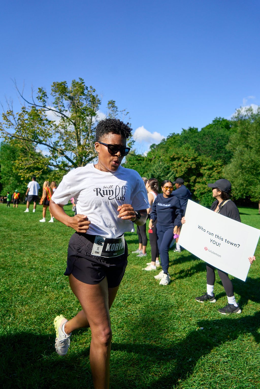 A woman running in a cross-country event on a grassy field with other participants and supporters, some holding signs. Bright sunny day with blue sky and green trees in the background.