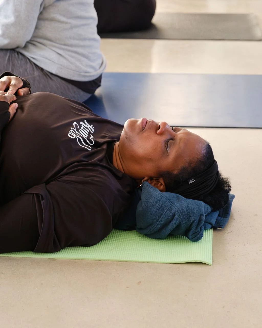 A person lying on a yoga mat with eyes closed, resting with a towel under their head in a gym or fitness studio.