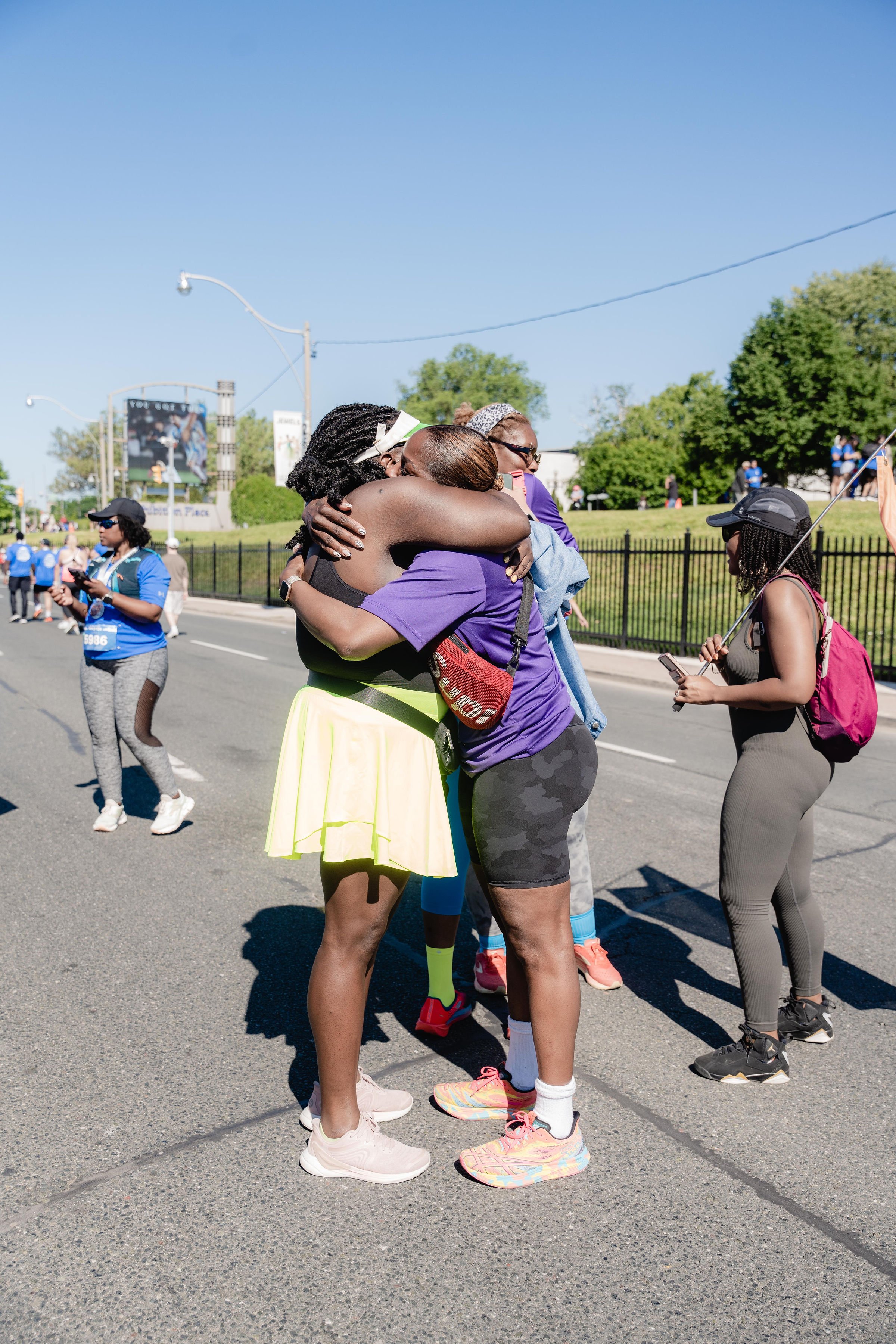 Two women hugging in the middle of a street during a marathon event, with other participants and spectators in the background on a sunny day.