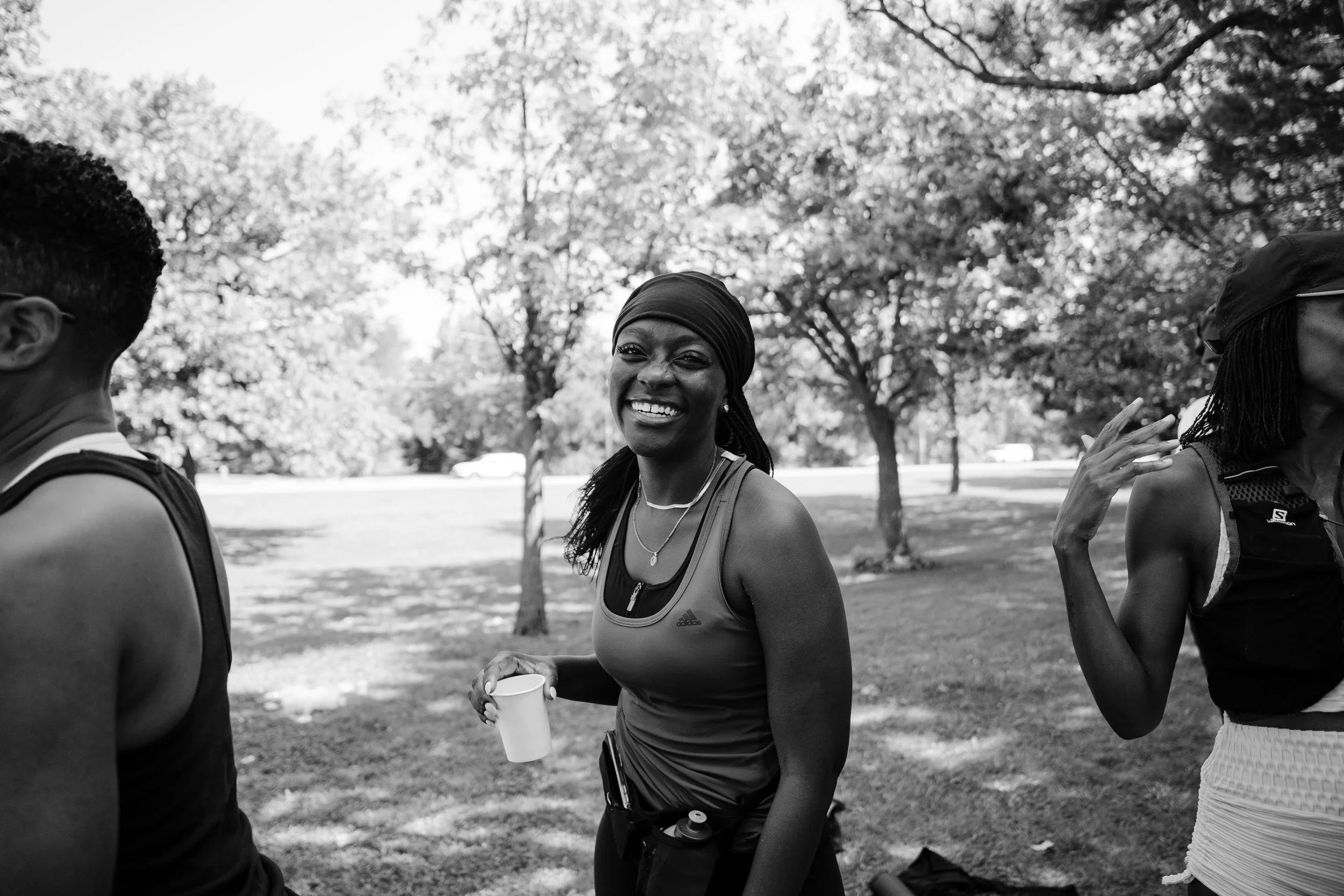 Smiling woman with headscarf holding a cup at an outdoor park, with trees in the background.