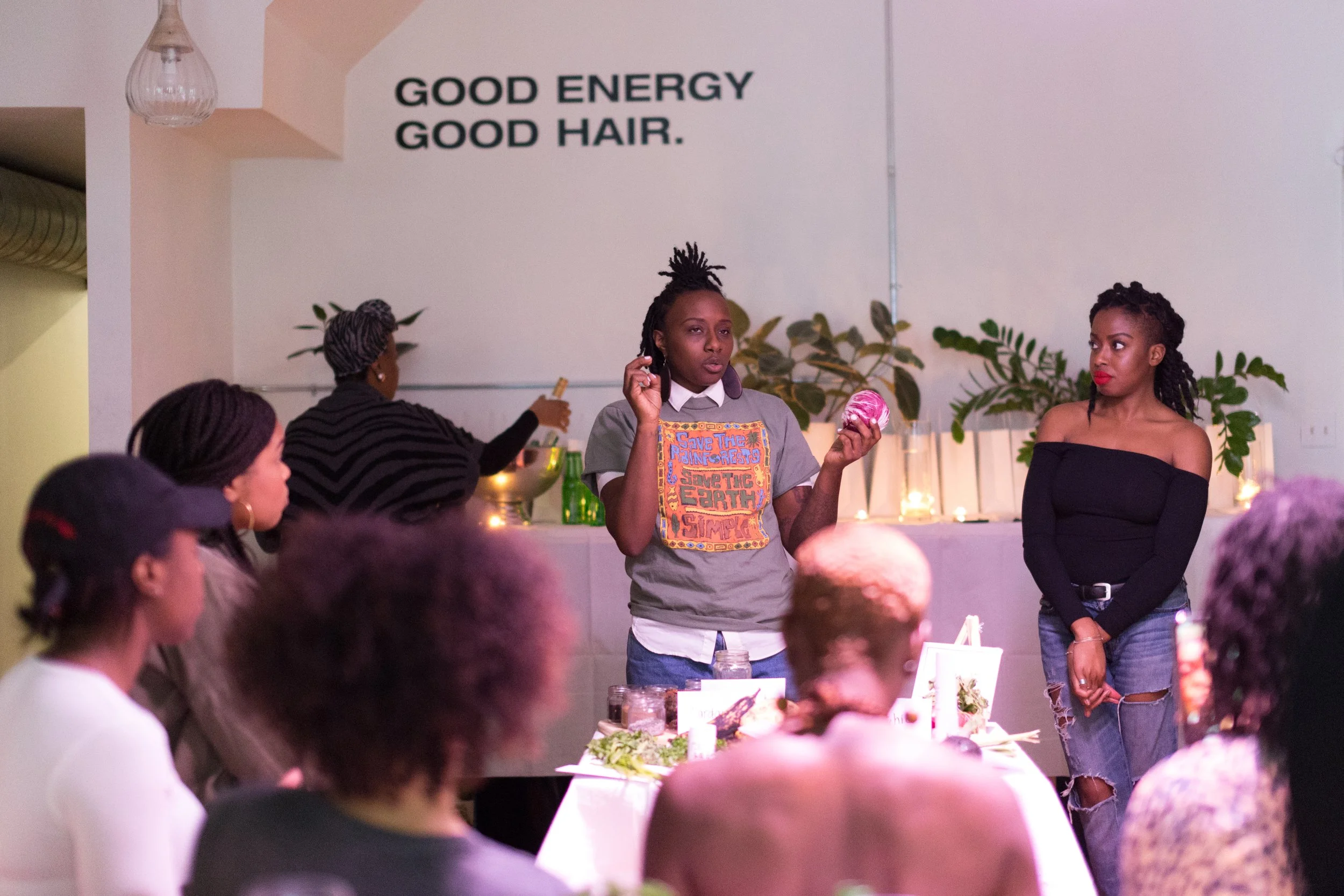 A woman with dreadlocks holding a small pink object and speaking to a group at a gathering, with a sign in the background that says 'Good Energy Good Hair' along with other women listening.
