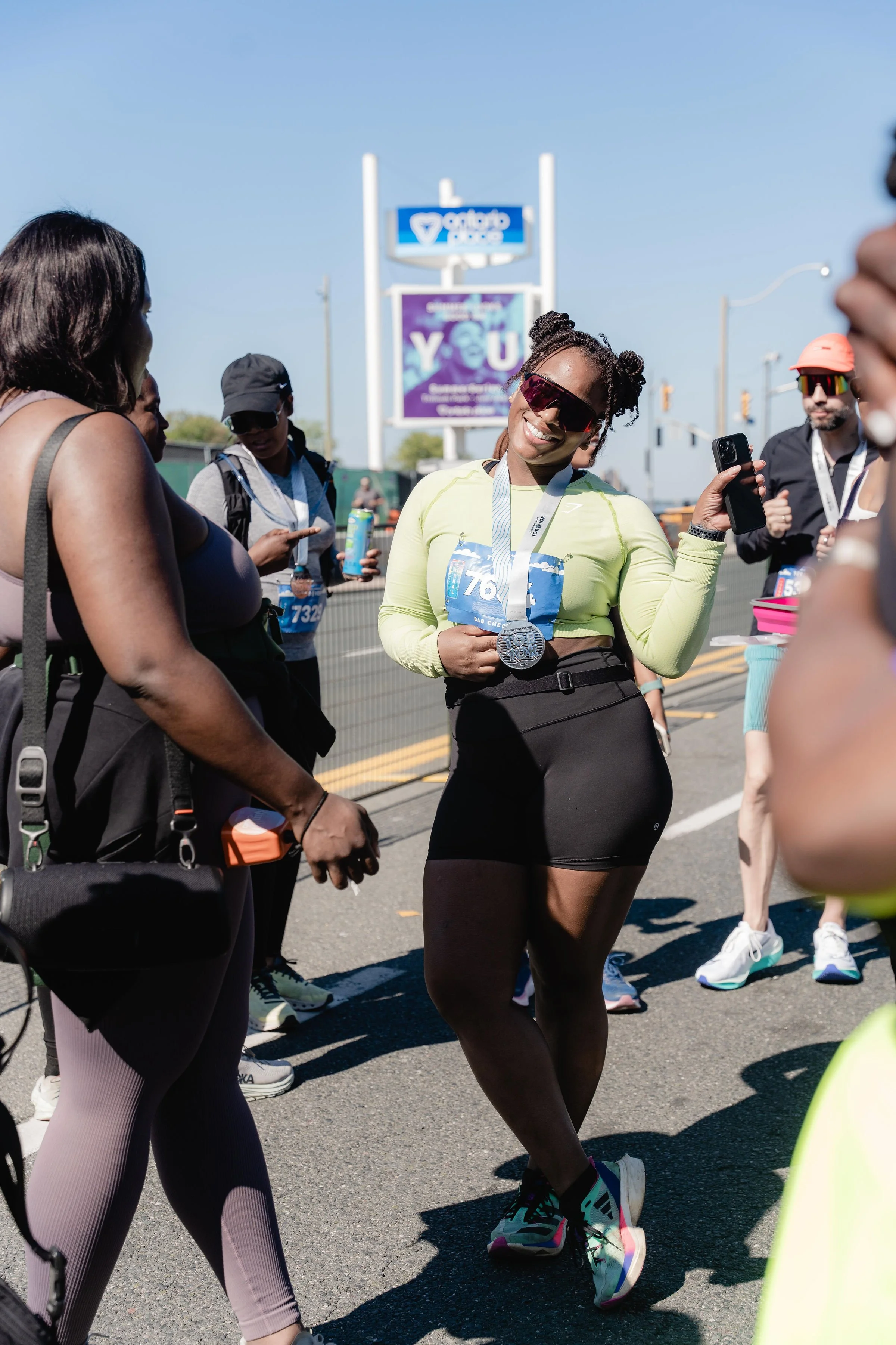 Woman with sunglasses and a marathon medal around her neck, smiling and holding a phone, after completing a race, surrounded by other runners or supporters on a sunny day.