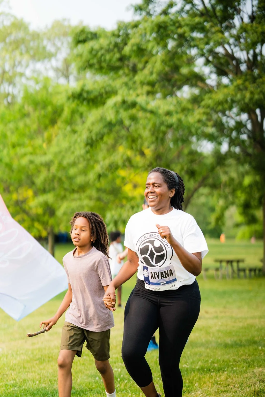 A woman and a young girl are running outdoors on grass with green trees in the background during daytime, smiling and holding hands.