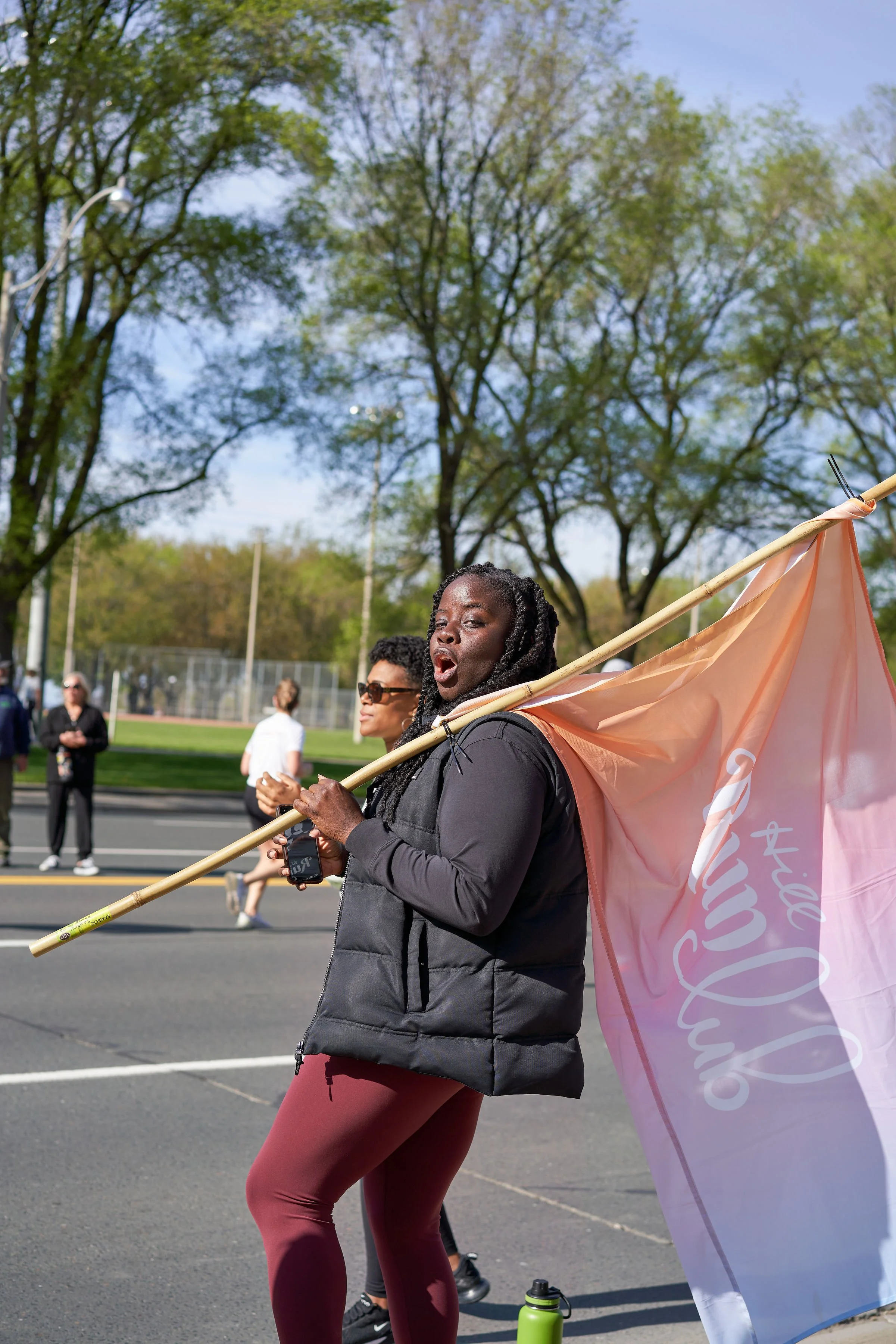 A woman holding an LGBTQ Pride flag on a parade street with trees and people in the background.