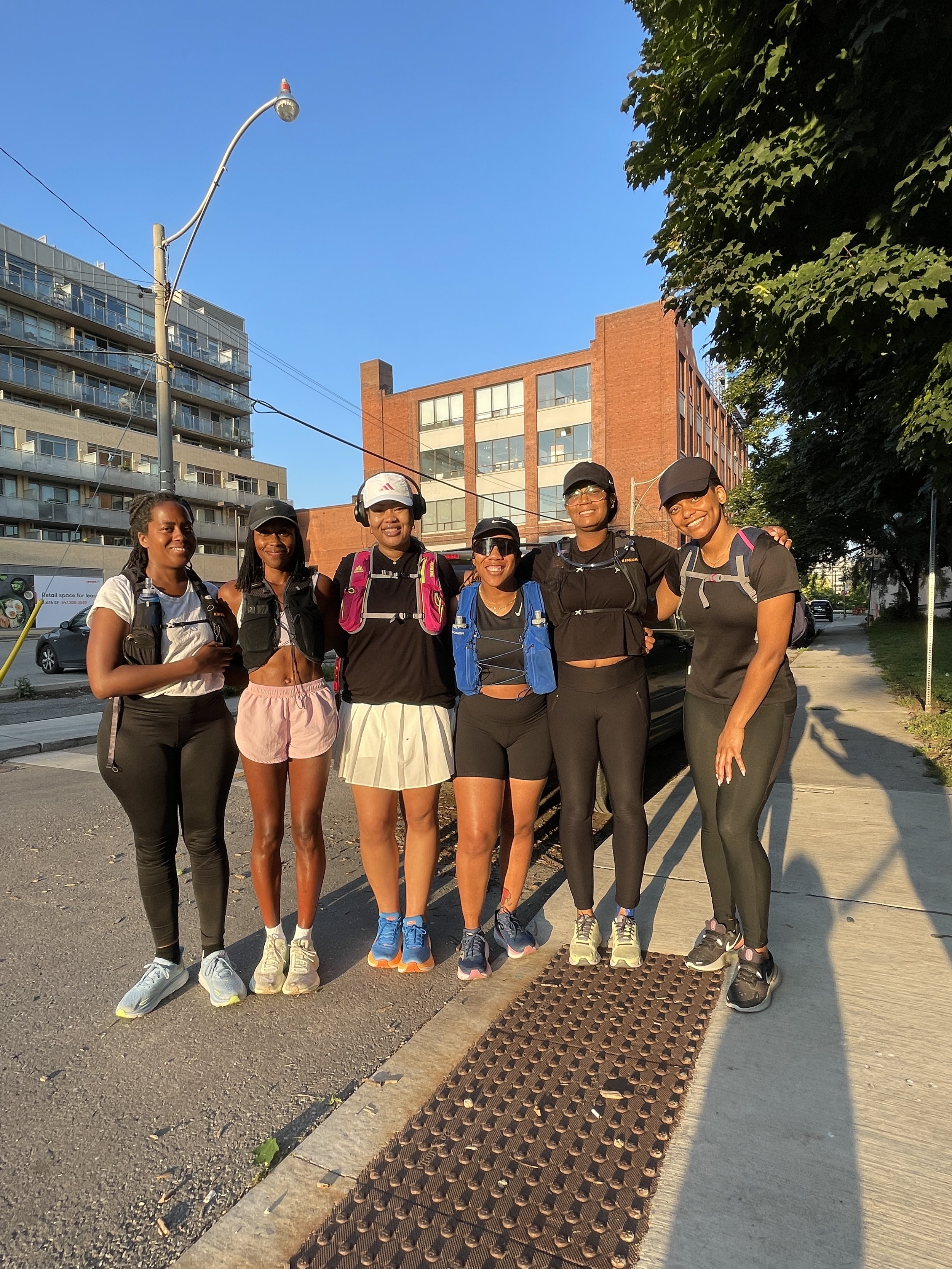 Group of six women in athletic wear standing together outdoors on a city sidewalk, smiling, with apartment buildings, trees, and a clear blue sky in the background.