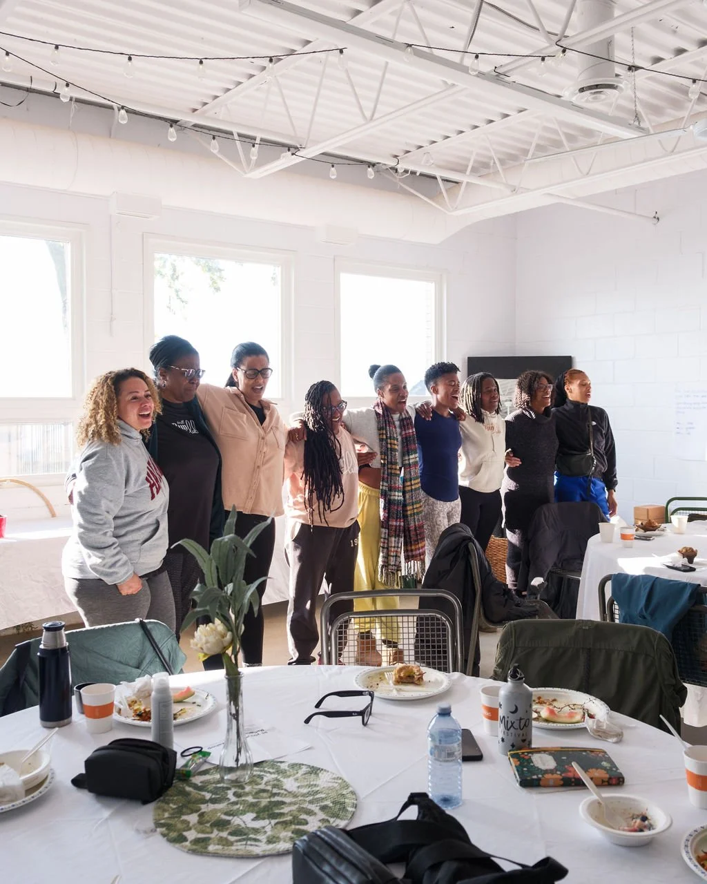 A group of women standing together with their arms around each other in a bright, white-walled room with large windows. They appear happy and are likely celebrating or concluding an event.